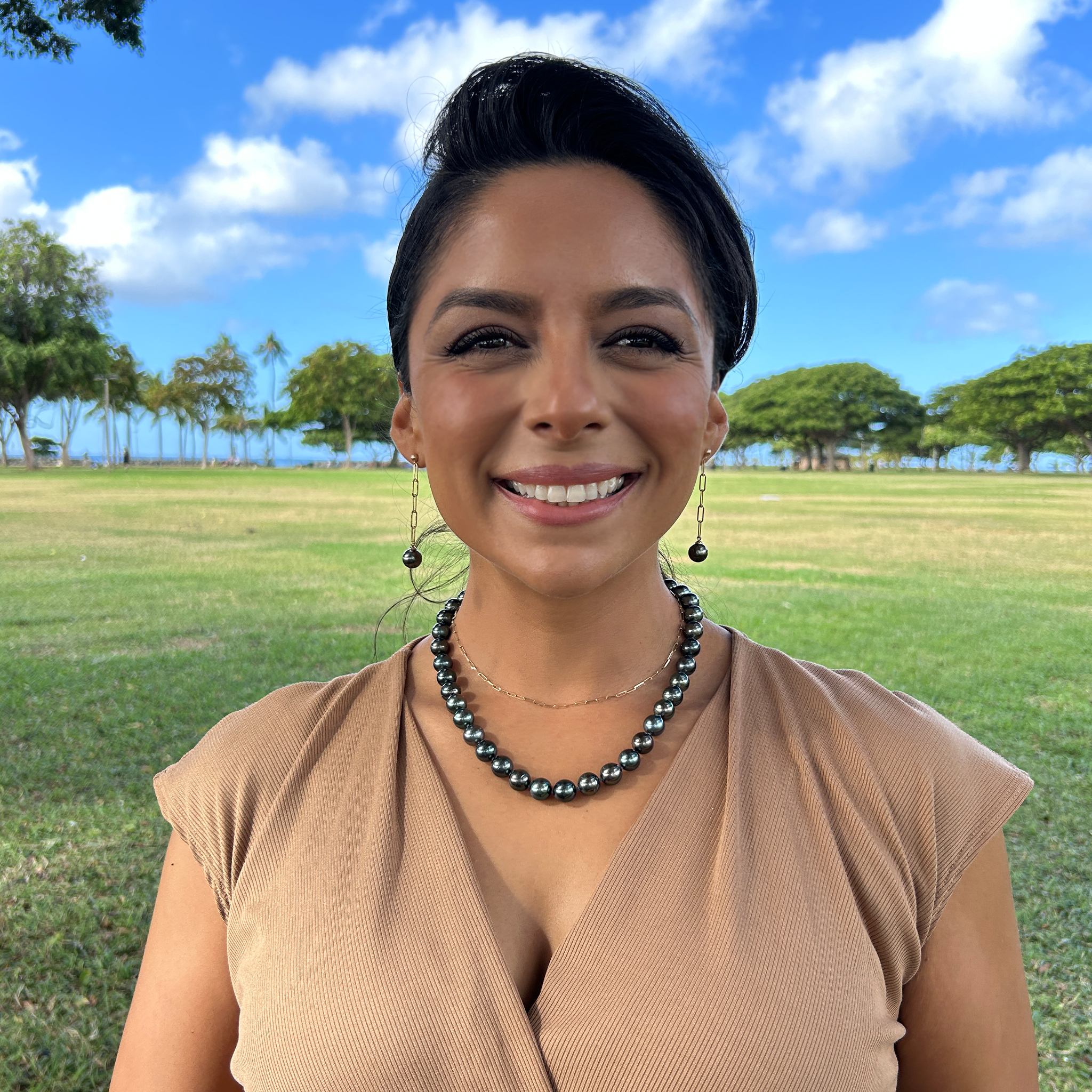 A woman in a brown top wearing a Tahitian black pearl strand and dangling Tahitian pearl earrings with a field and trees in the background