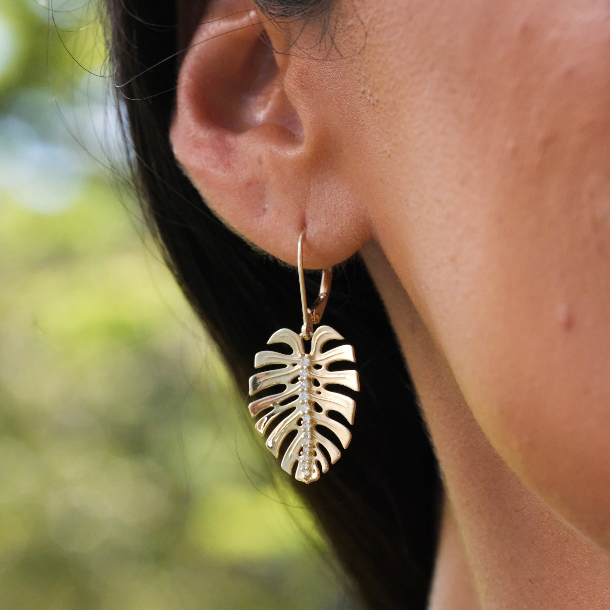 A woman's ear with 23mm Monstera Earrings in Gold with Diamonds against a blurred green background