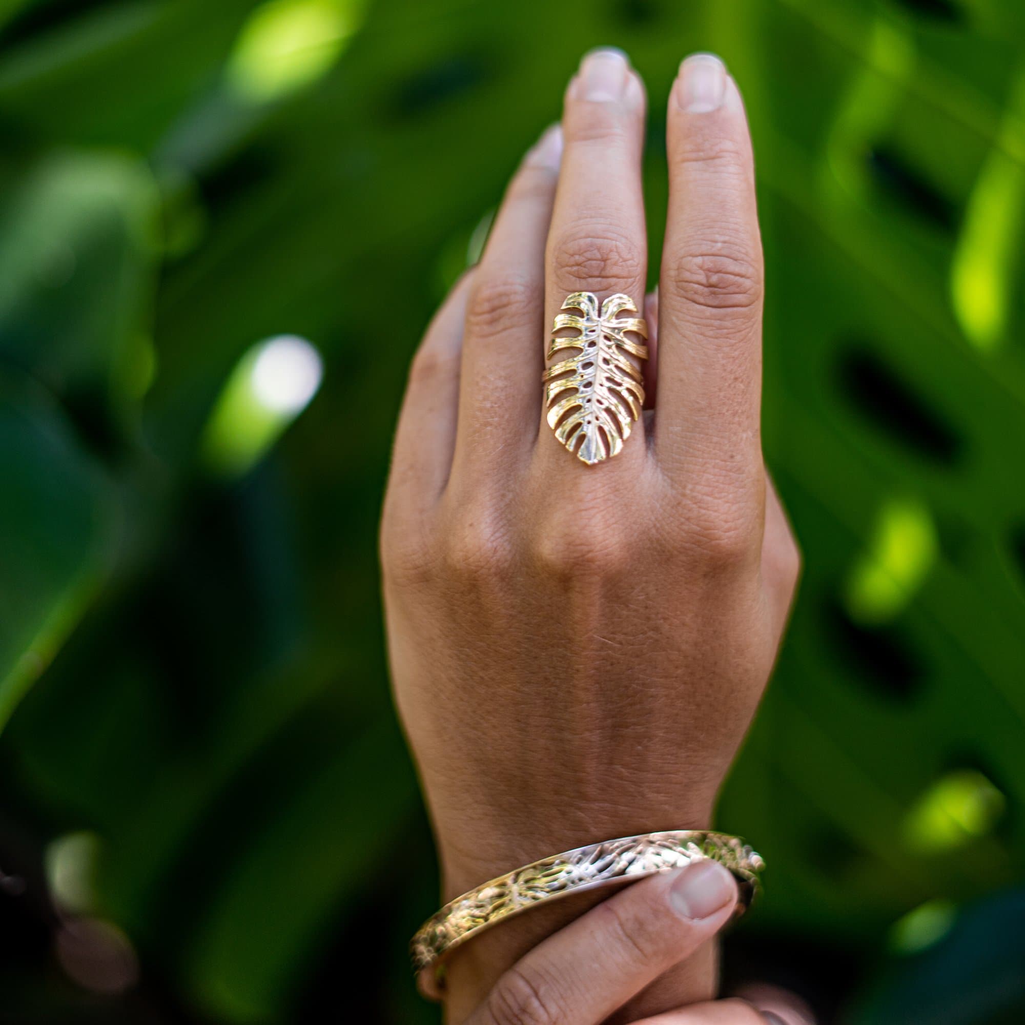Close up of a 30mm Monstera Ring in Gold on a hand with a blurred monstera leaf in the background