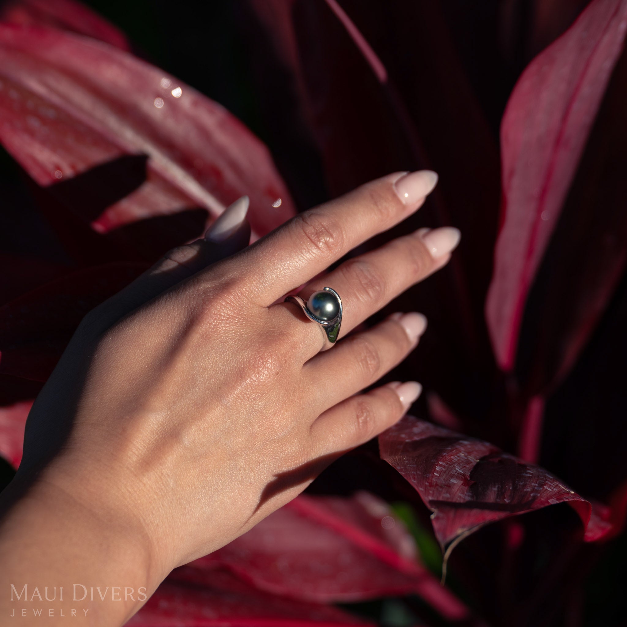 Hand wearing a ring with a Tahitian black pearl against a red leaf background