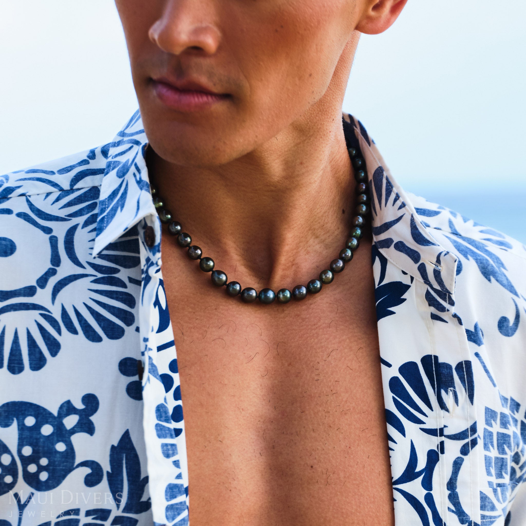 Man wearing a blue and white patterned shirt with a Tahitian black pearl strand, against a blurred natural background.