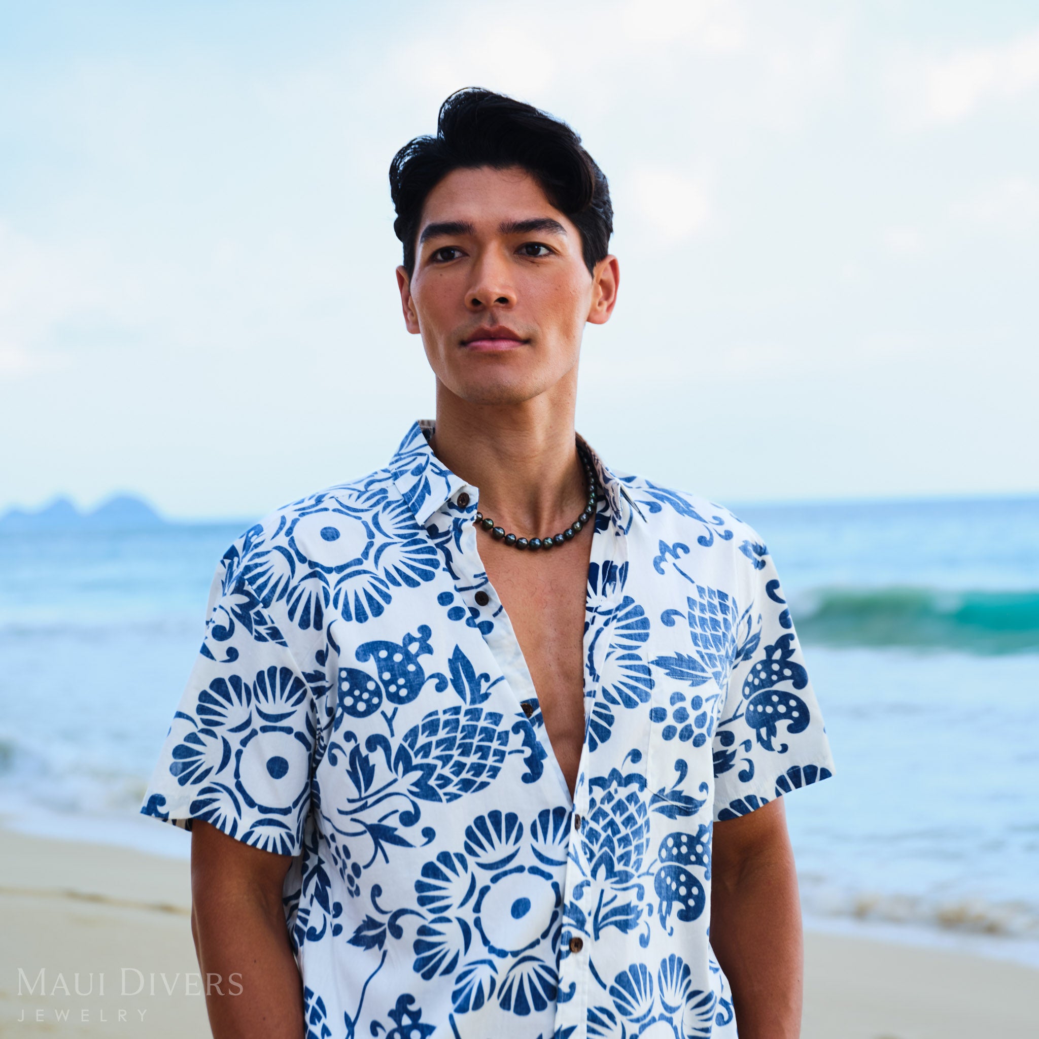 Man wearing a blue and white patterned shirt with a Tahitian black pearl strand, against a blurred beach background.
