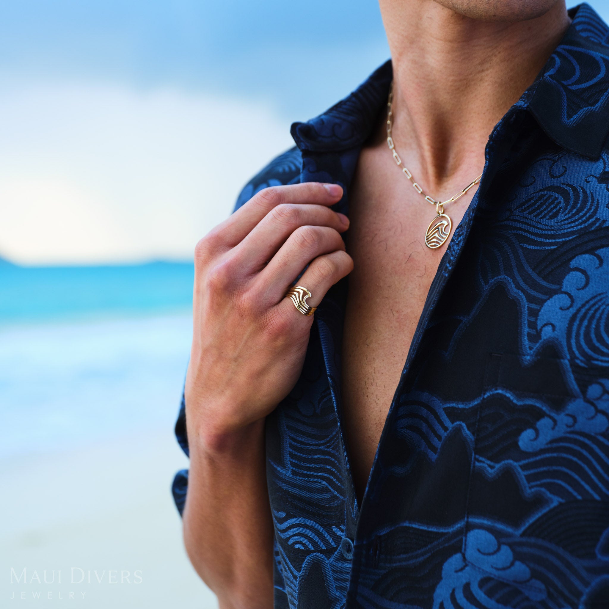 Man wearing a blue patterned shirt with a Modern style wave necklace and ring, against a blurred beach background.