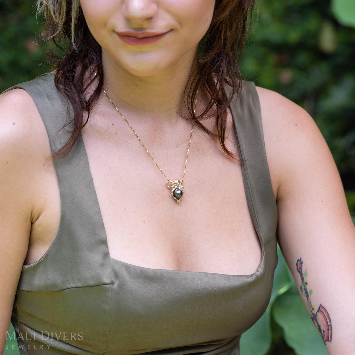 A close-up lifestyle shot of a woman wearing a gold octopus pendant with a Tahitian black pearl wrapped in tentacles on a glistening box chain, styled with an olive green satin top. 