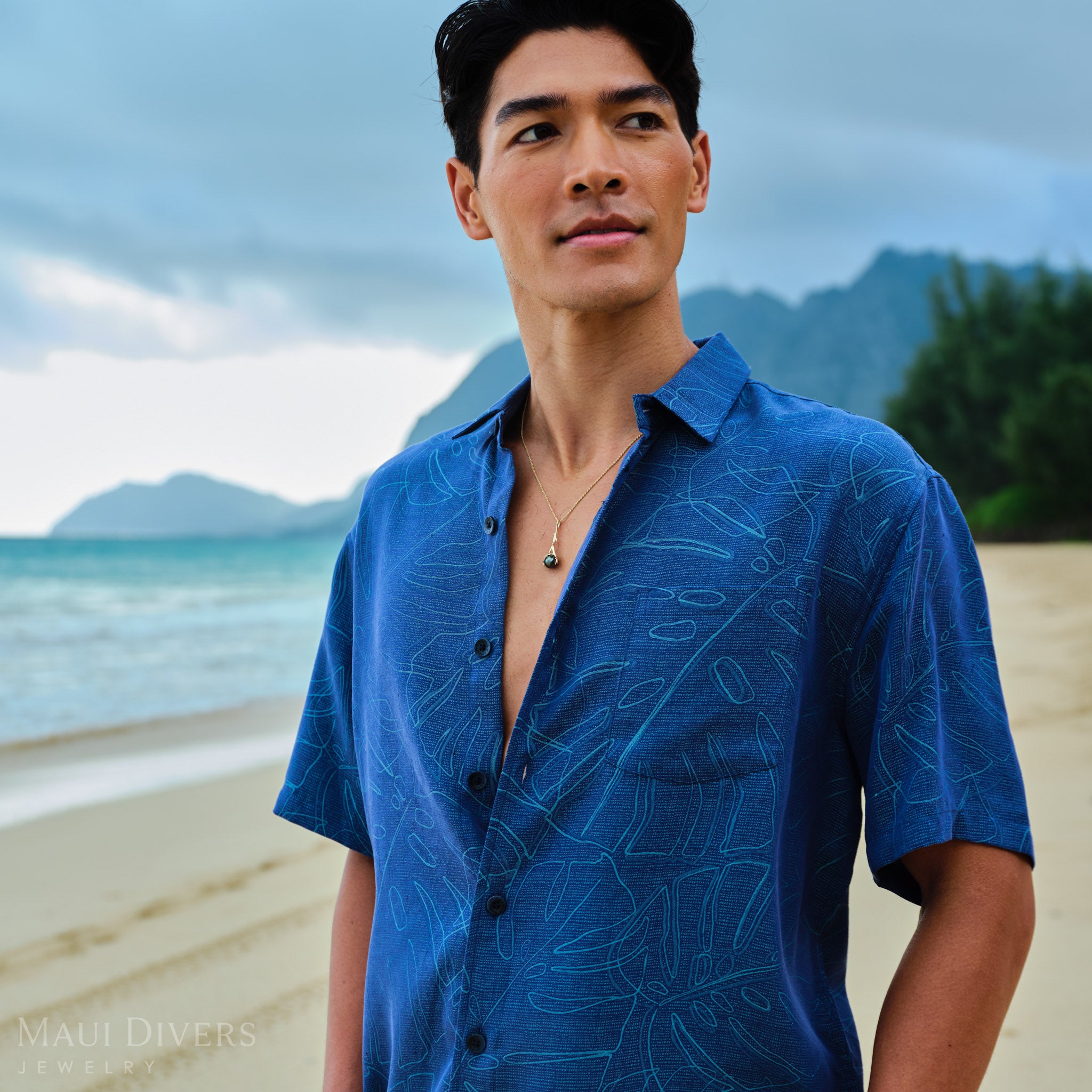 Man wearing gold Tahitian black pearl pendant a blue patterned shirt on a beach with mountains in the background