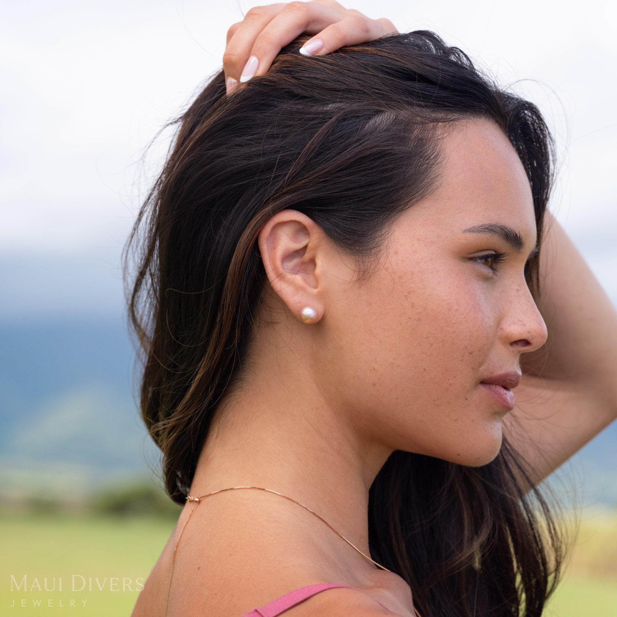 Side view of a woman wearing a freshwater pearl natural color stud earring in 14k yellow gold as she runs her fingers through her hair with a blurry green and blue background