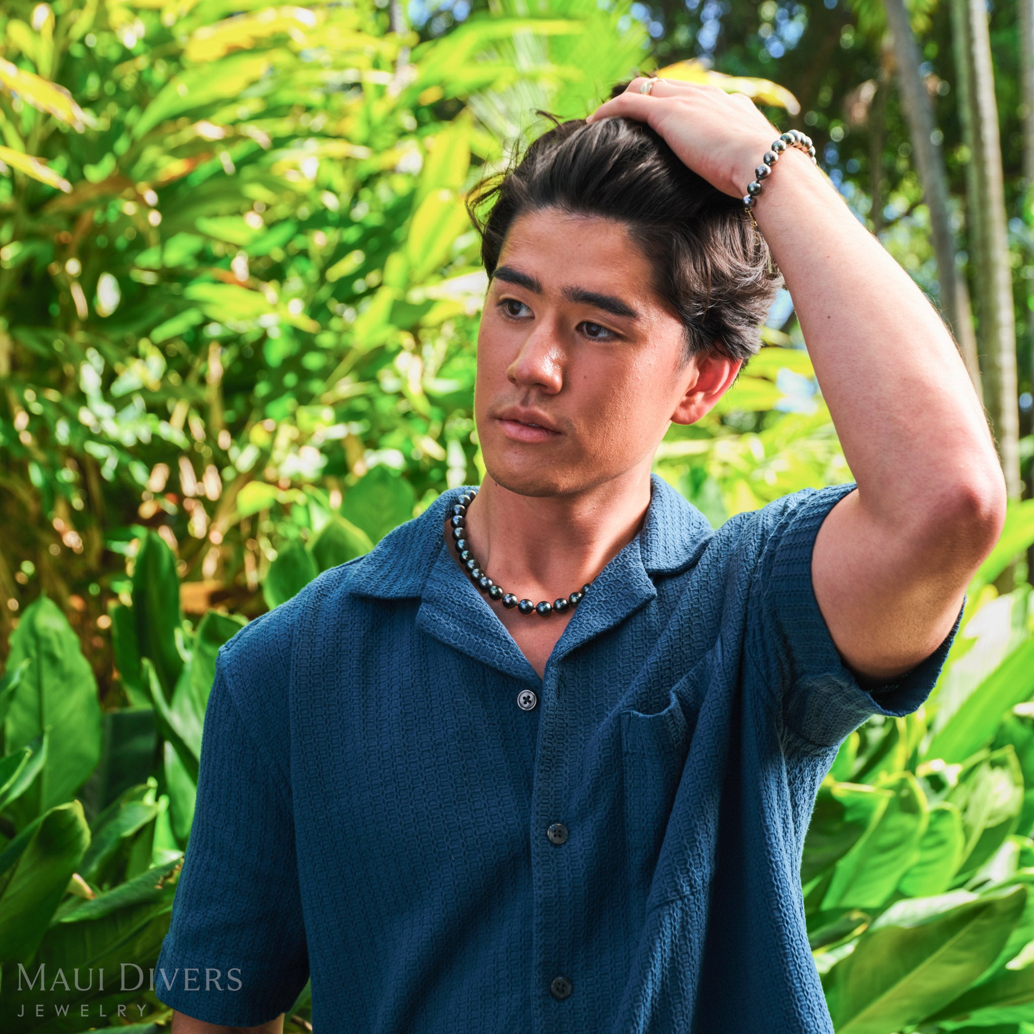 Man in a blue shirt wearing a Tahitian black pearl strand necklace with a matching Tahitian black pearl bracelet, against a blurred forest background