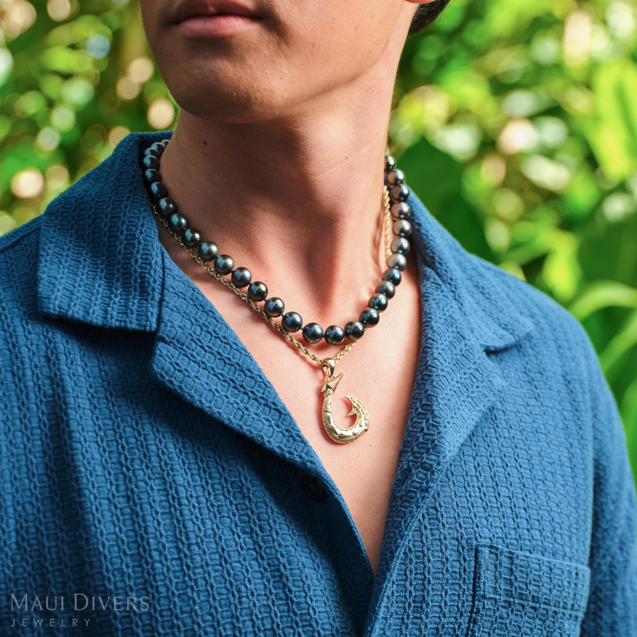 Close-up of a man in a blue shirt wearing a Fish Hook Pendant in 14k yellow gold, paired with a single Tahitian black pearl strand, against a blurred leafy green background
