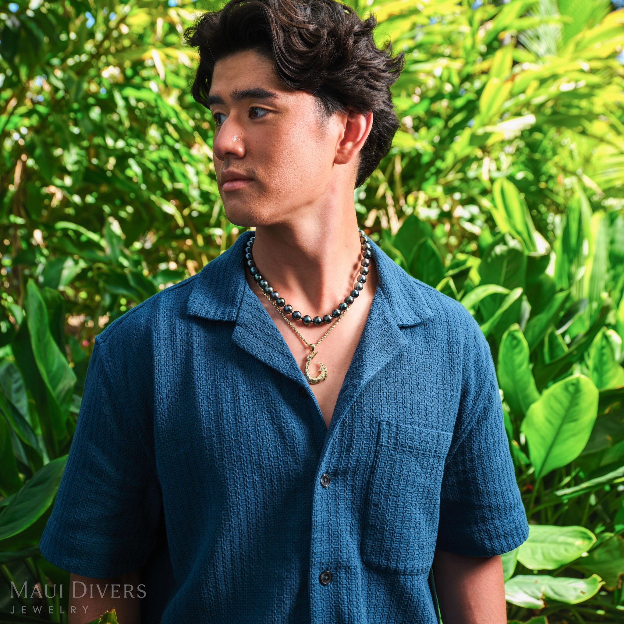Man in a blue shirt wearing a Fish Hook Pendant in 14k yellow gold, paired with a single Tahitian black pearl strand, against a blurred leafy green background