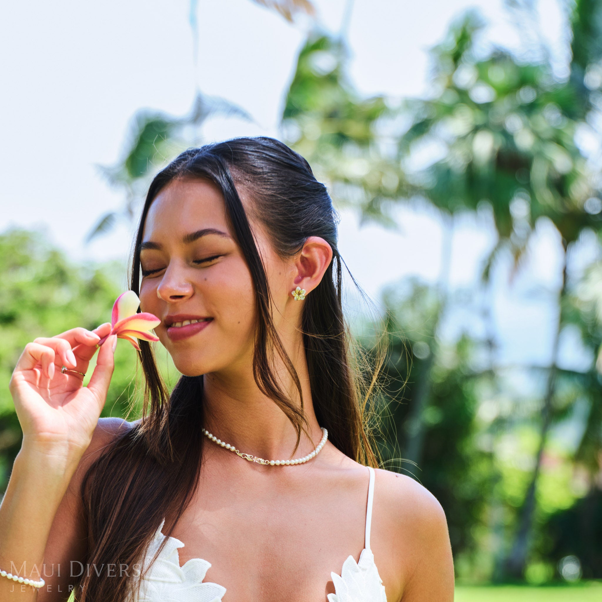 Smiling woman smelling a plumeria flower wearing a Hawaiian Heirloom Plumeria Freshwater White Pearl Necklace in Two Tone Gold with Diamond against a blurred park background