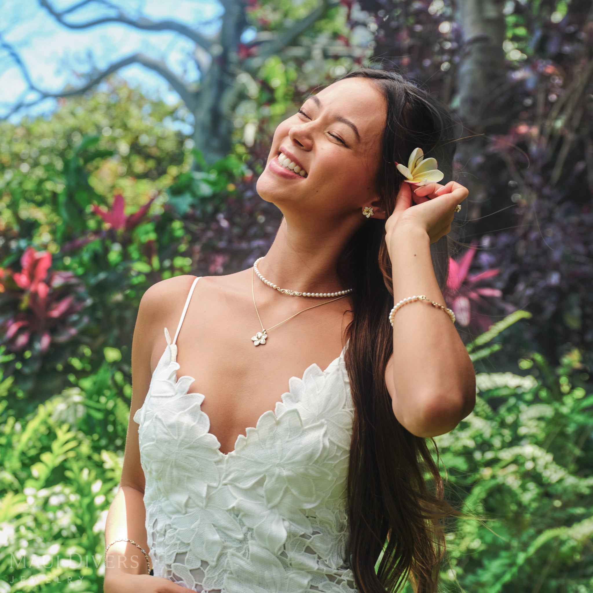 Smiling woman with a plumeria flower behind her ear wearing Plumeria Mother of Pearl earring and pendant in 14k yellow gold and a Hawaiian Heirloom freshwater pearl strand necklace against a blurred tropical background