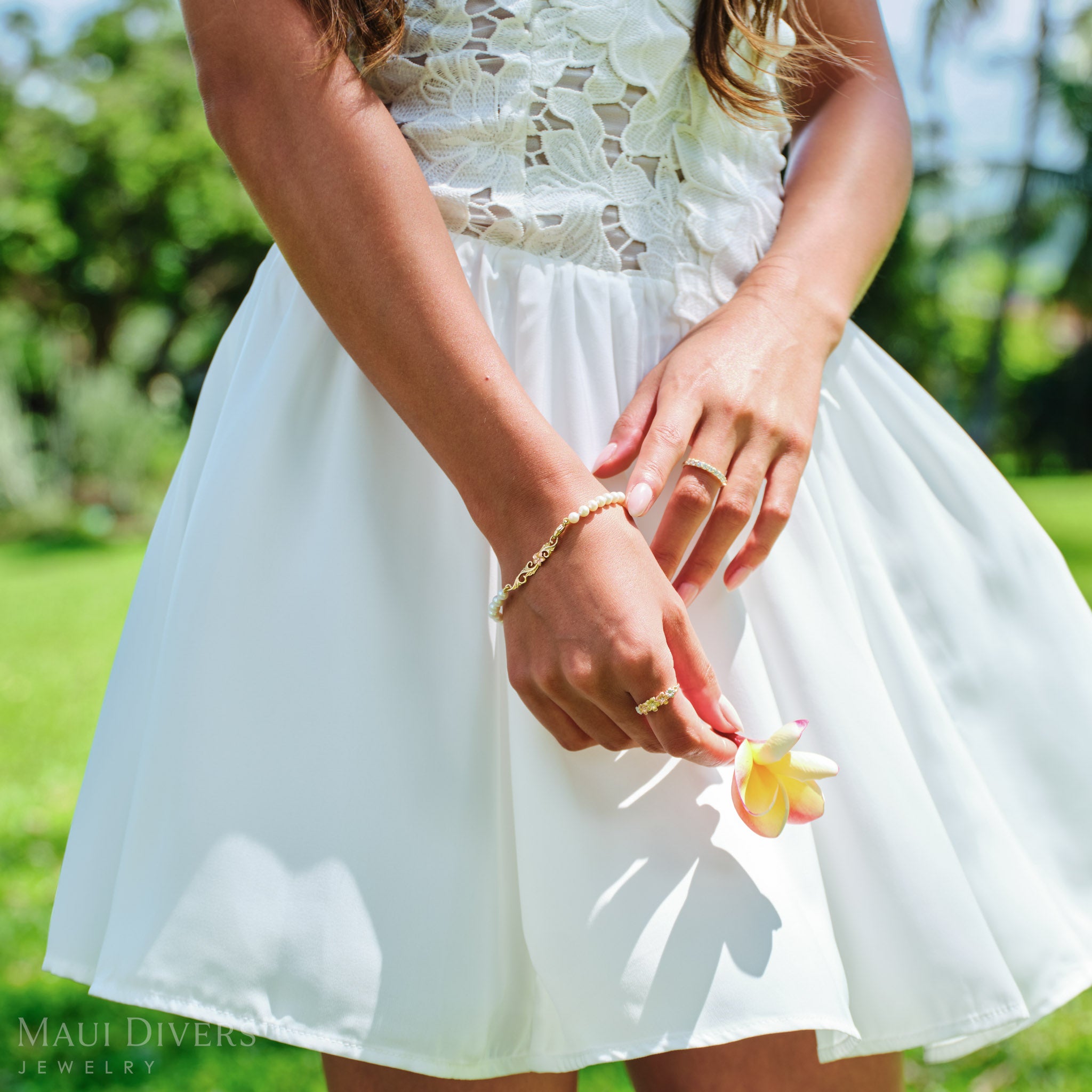 Woman in a white dress wearing a Hawaiian Heirloom Plumeria Freshwater White Pearl Bracelet in two tone gold with diamond and a Plumeria Tri Color Ring, holding a plumeria flower against a blurred forest background