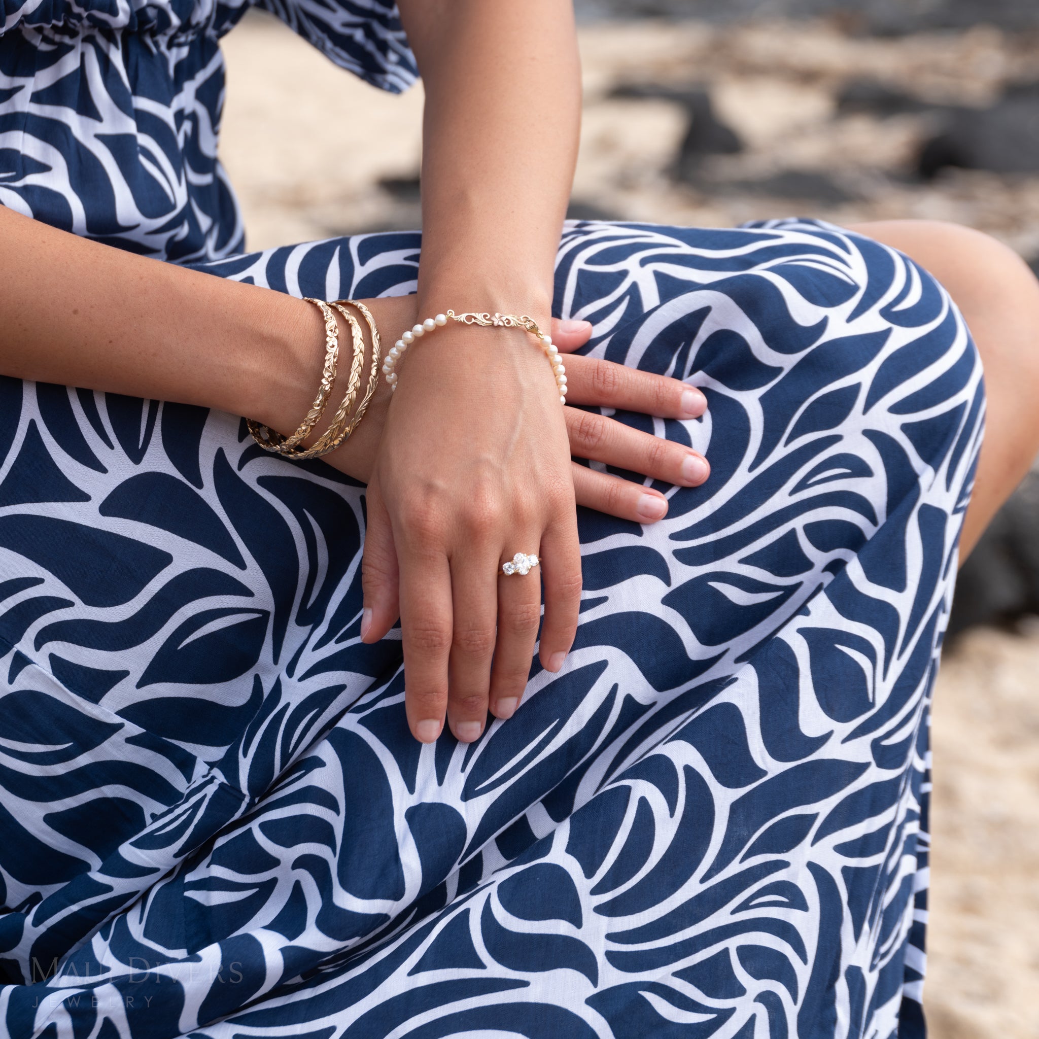Person wearing a blue and white patterned dress, 3-stone Hidden Hawaiʻi lab-grown diamond ring, Hawaiian Heirloom pearl bracelet, and Hawaiian Heirloom bangles against a blurred natural background