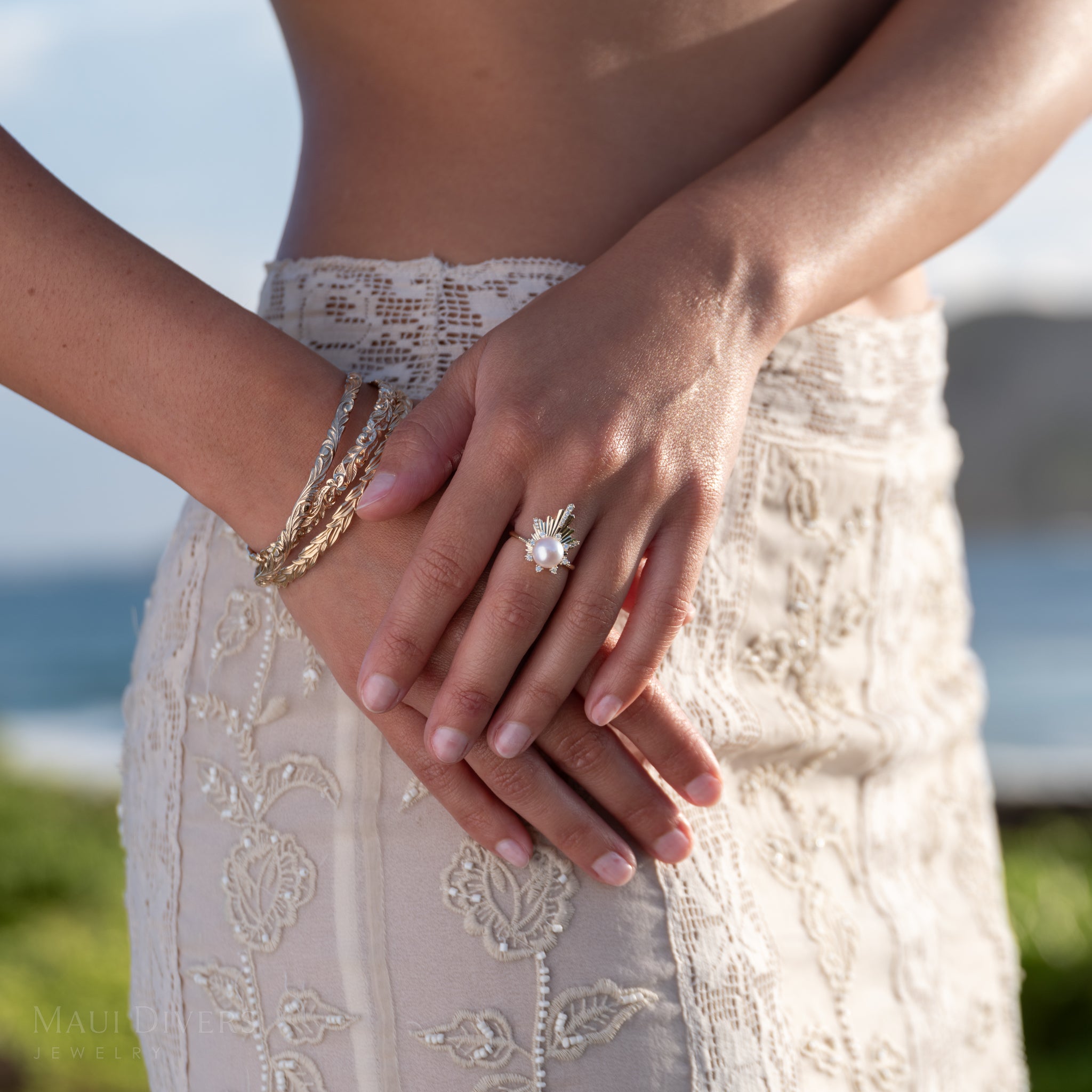 Close-up of hands wearing Hawaiian bracelets and the E Hoʻāla Akoya White Pearl and diamond Ring in yellow Gold with a scenic background