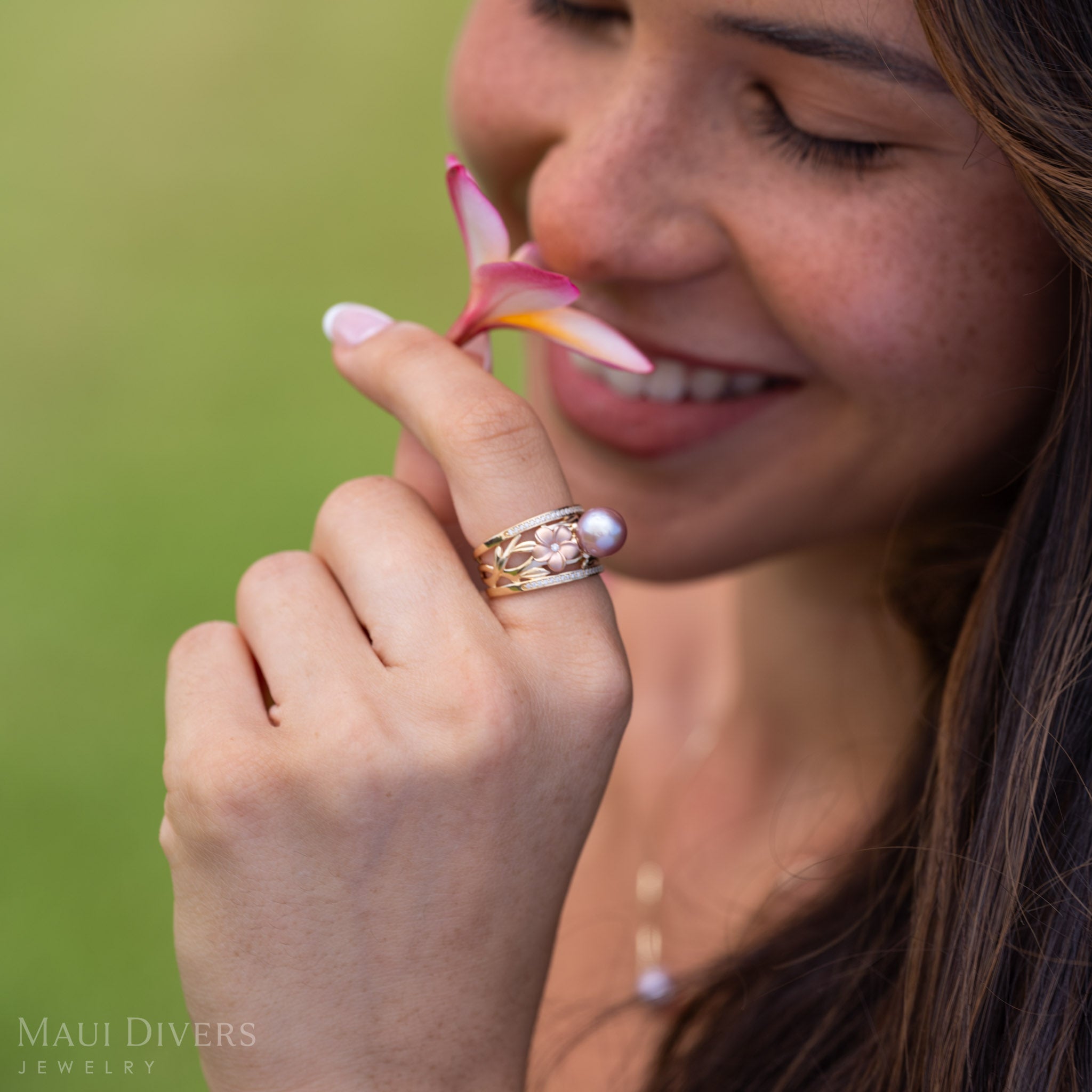 A smiling woman wearing a Natural Lavender Freshwater Pearl Ring with Tri Color Plumerias in 14k Gold accented with  Diamonds smelling a light pink and yellow plumeria flower.
