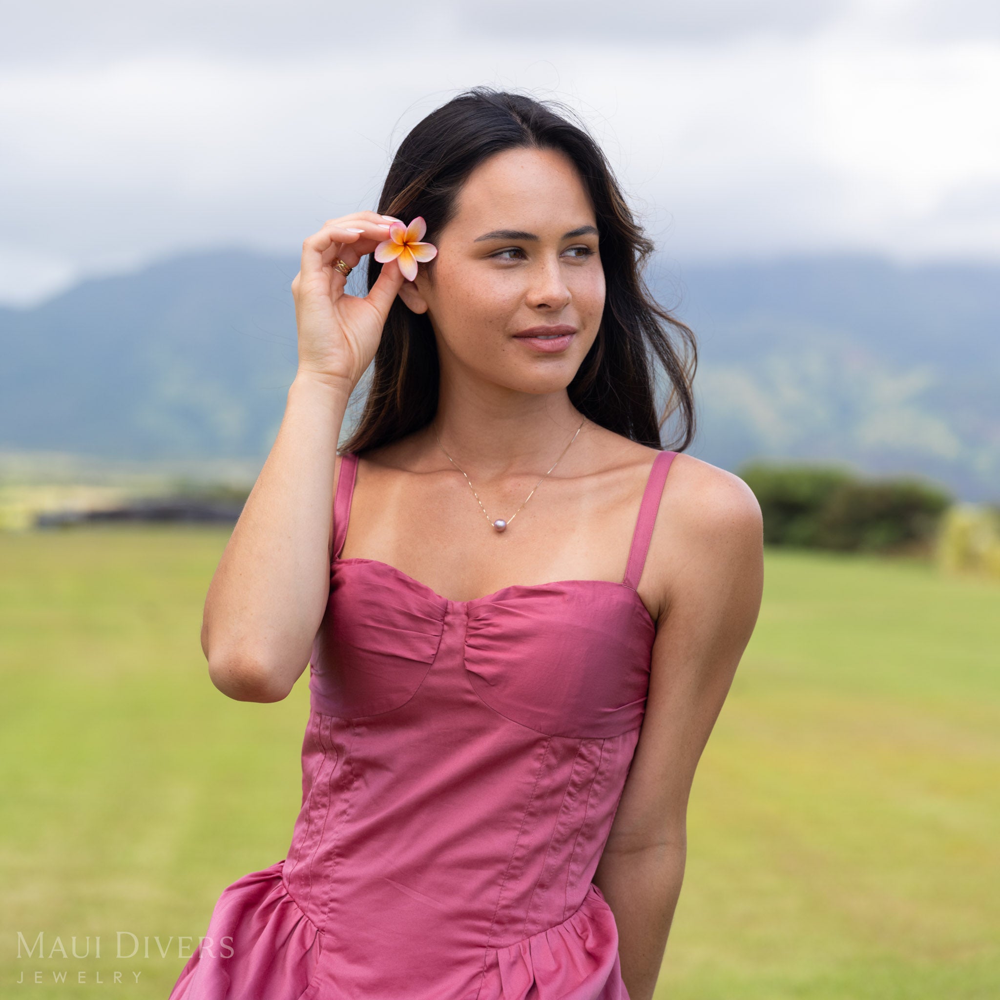 Smiling woman in a pink dress wearing a Freshwater Lilac Pearl Necklace in 14k yellow gold around her neck, with a plumeria in her hair, against a blurred open field background