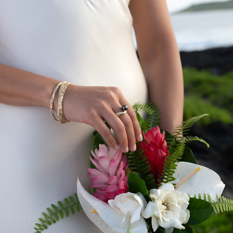Bouquet of flowers held by a person wearing a gold Kupukupu Fern Tahitian black pearl ring and Hawaiian bracelets against a white dress.