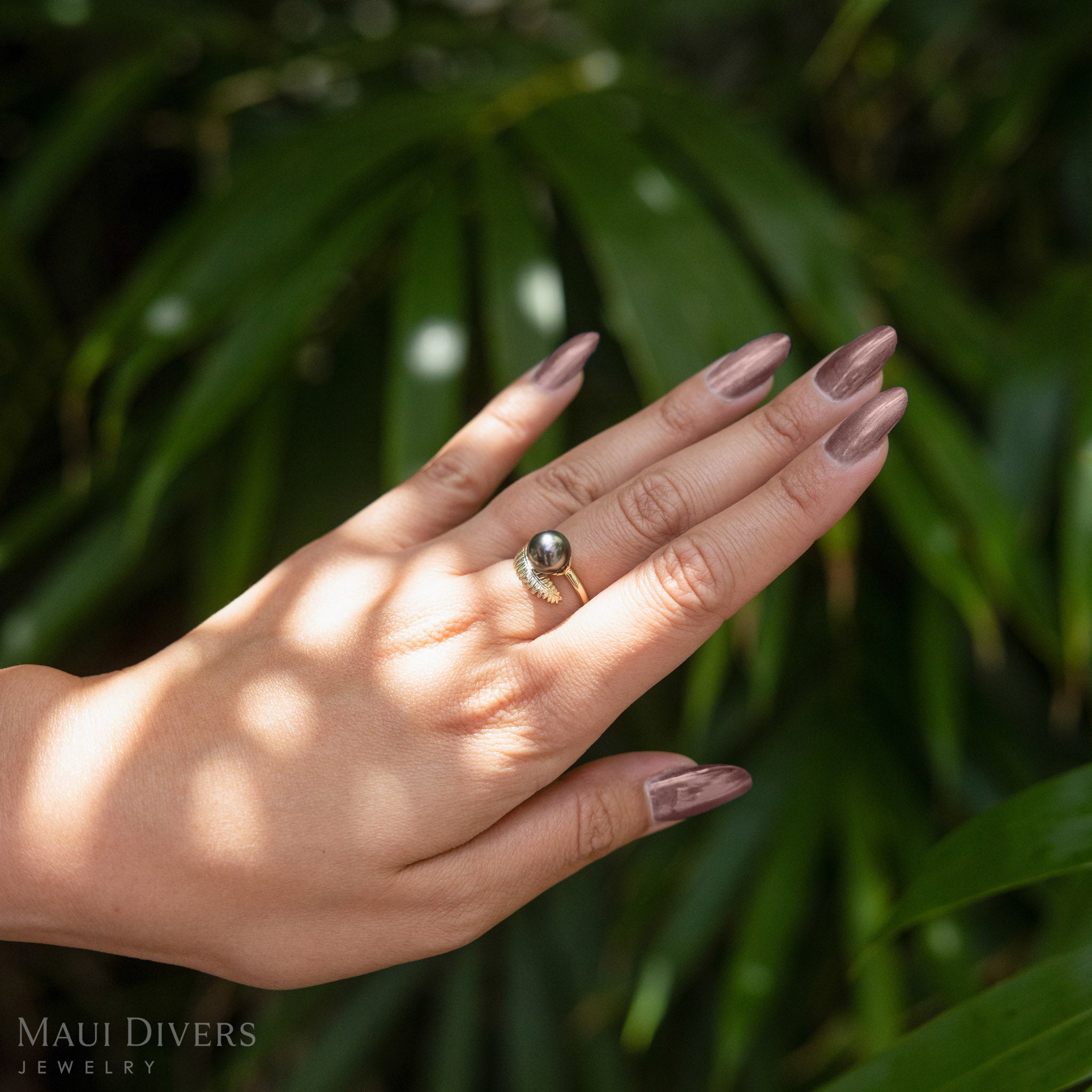 Hand wearing a kupukupu fern Tahitian black pearl ring against a green leafy background