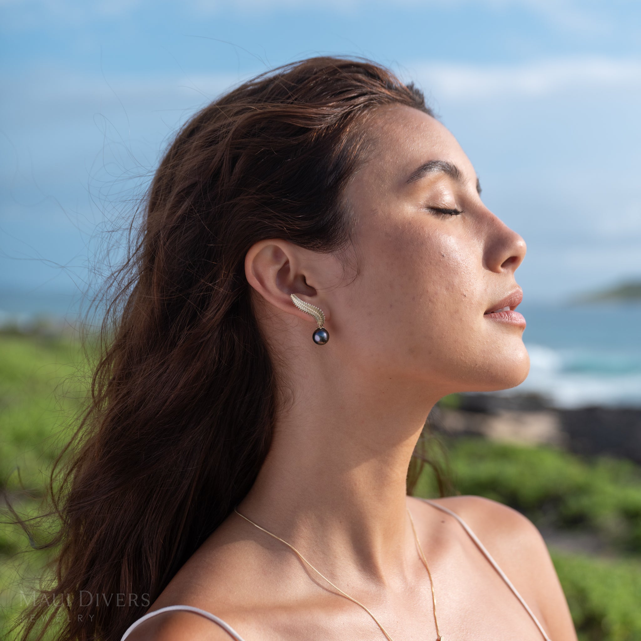 Close-up of a woman wearing the gold Kupukupu fern Tahitian black pearl climber style earrings with a scenic beach background