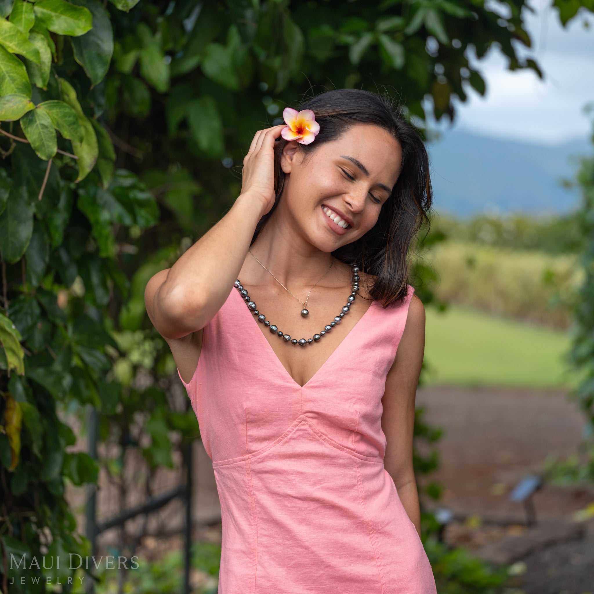 A smiling woman with a plumeria in her hair wearing a light pink dress with a Tahitian Black Pearl Pendant in 14k yellow gold, paired with a Tahitian Black Pearl Strand, against a blurred leafy green background