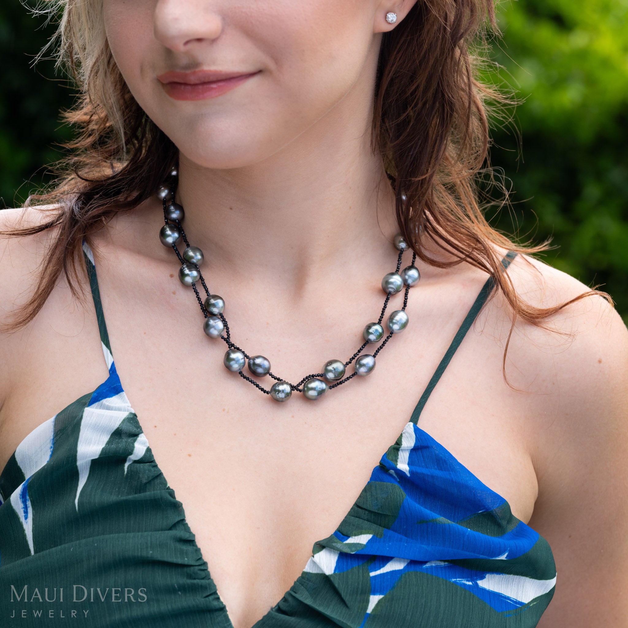 A close-up lifestyle shot of a woman wearing a double-layered baroque Tahitian black pearl and black spinel necklace with green foliage in the background. 