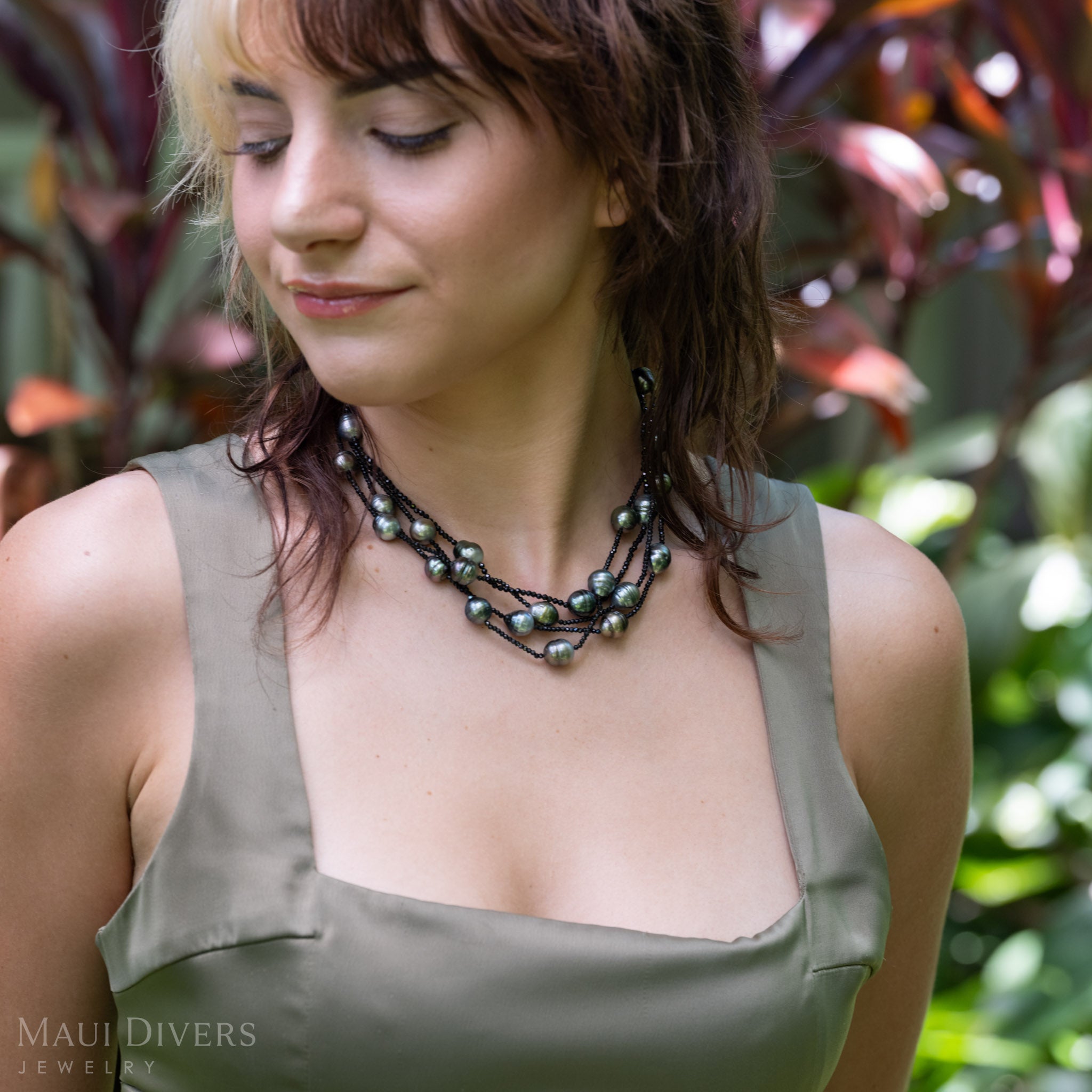 A woman wearing a multi-layered Tahitian black pearl and black spinel necklace with greenish-gray pearls, styled with an olive-green satin top against a blurred tropical foliage background. 
