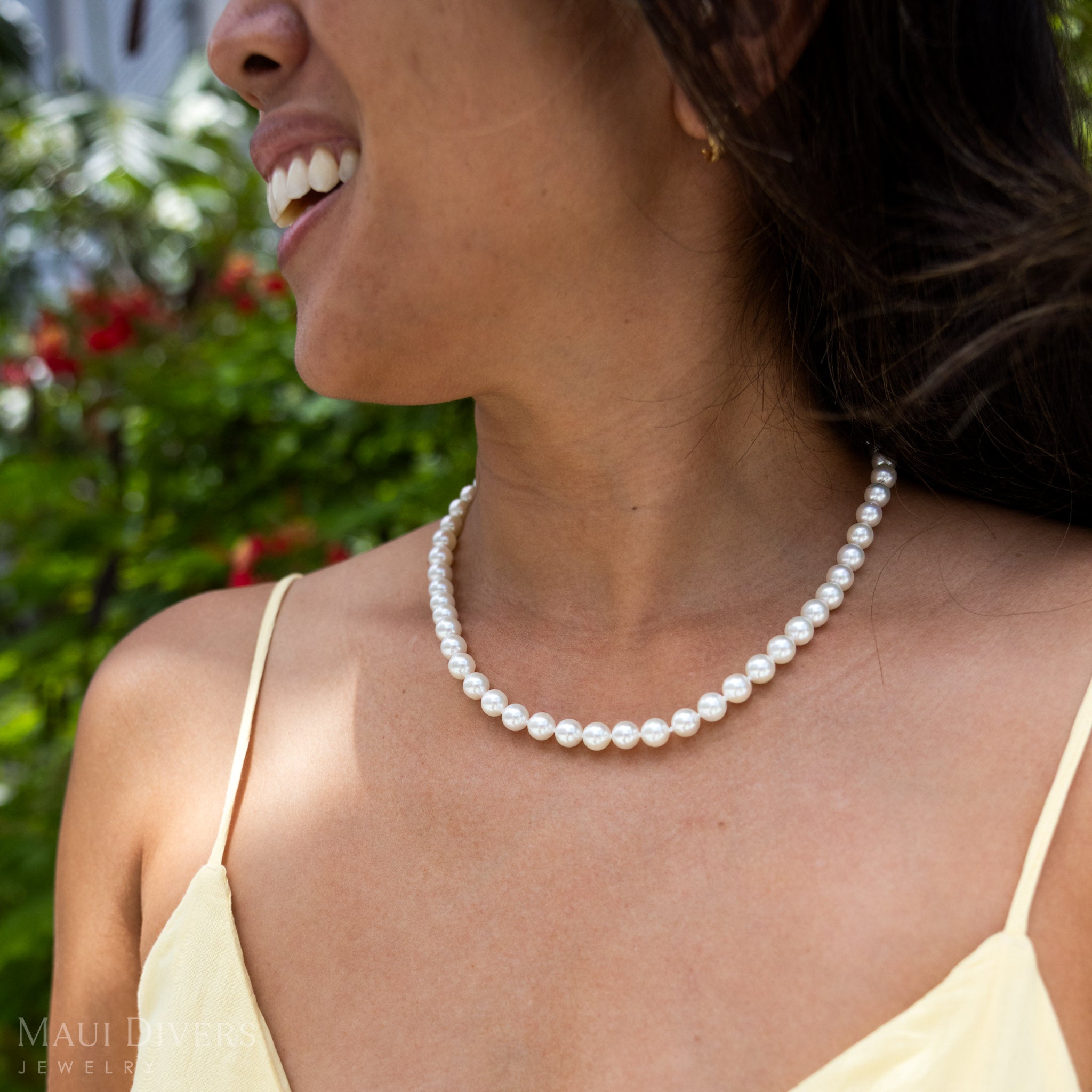 Smiling woman in a yellow top wearing an Akoya White Pearl Strand against a blurred leafy green background