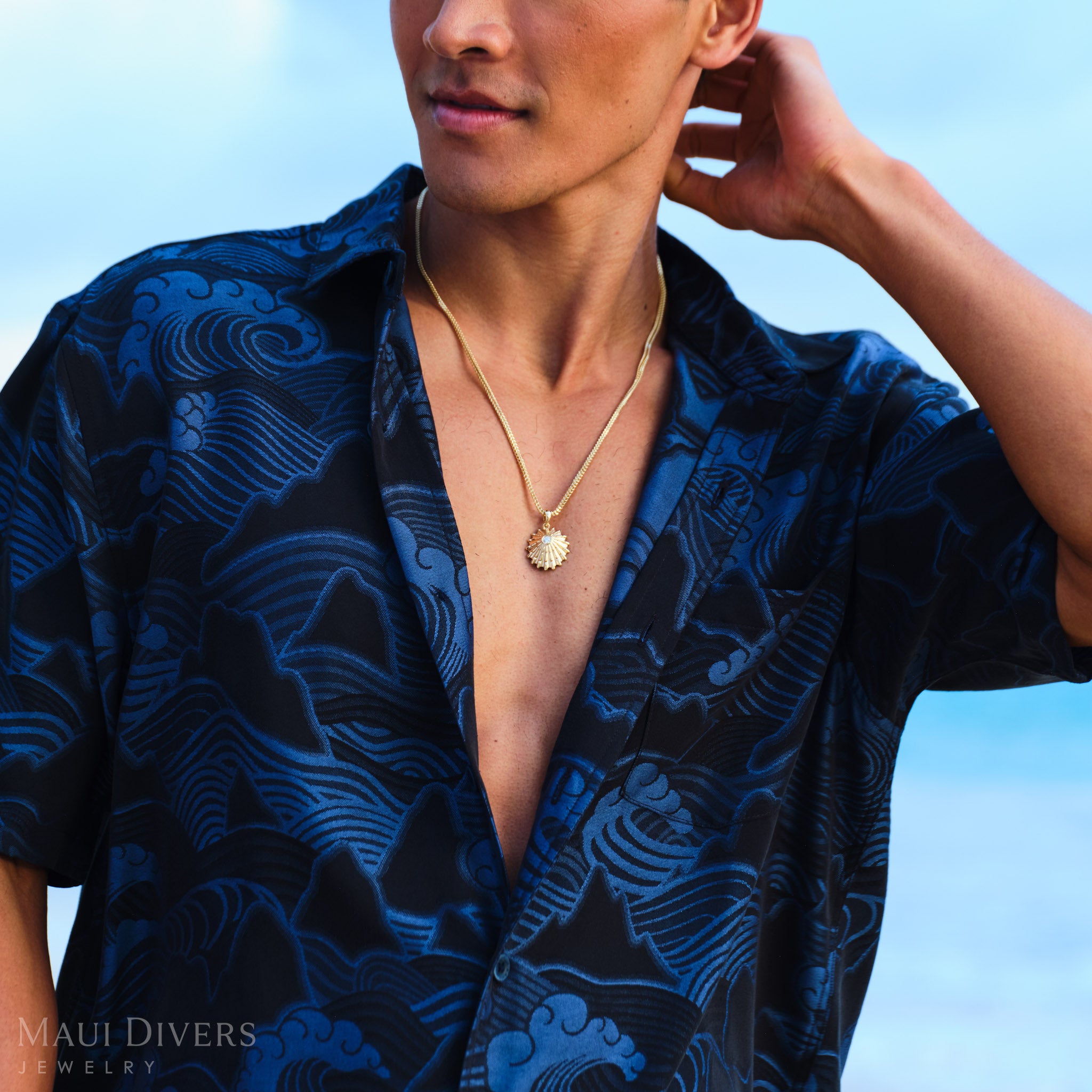 Man wearing a blue patterned shirt with a gold ʻopihi necklace against a clear sky.
