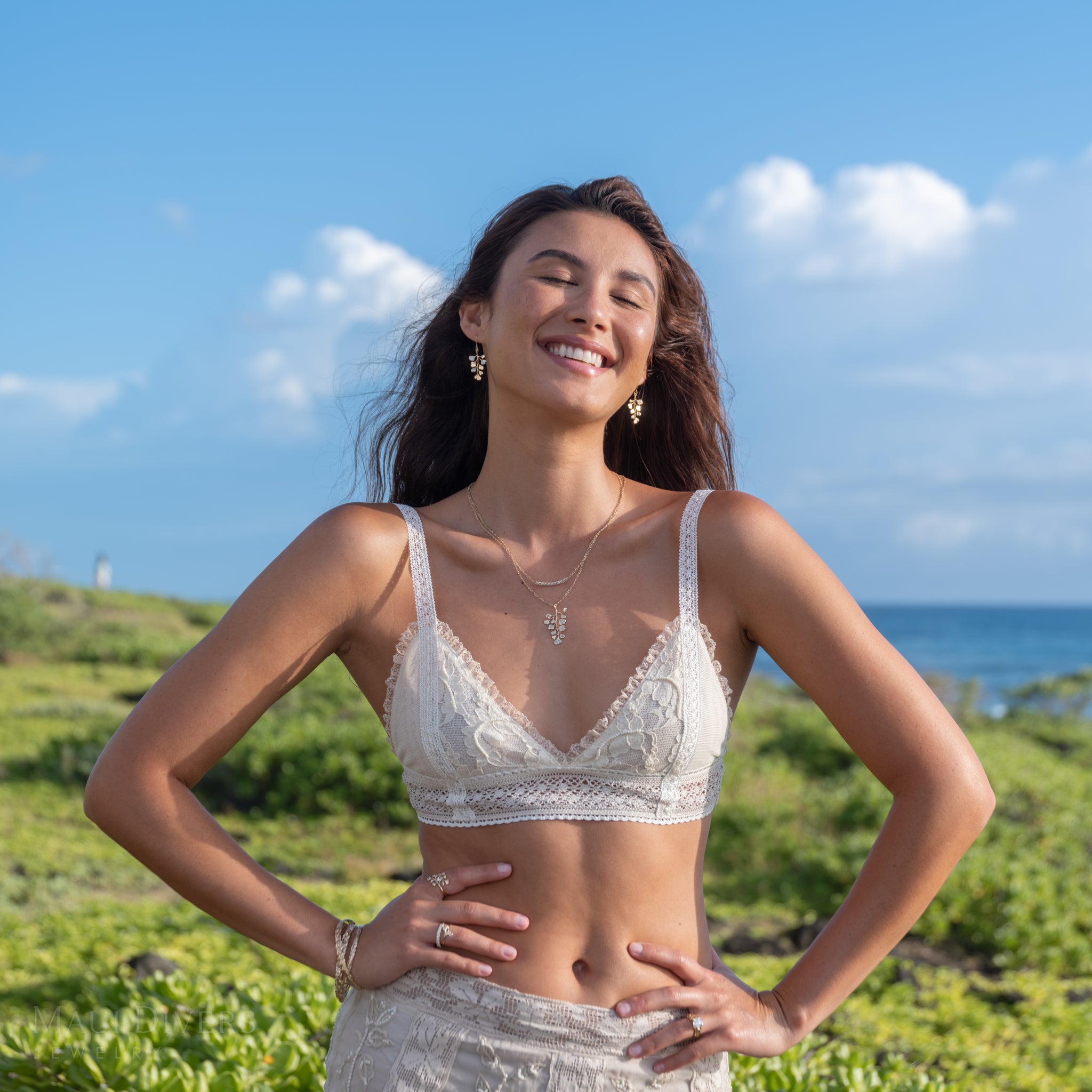 A woman stands outdoors on a grassy coastal hill wearing 14k yellow gold Maiden Hair Fern Earrings, Pendant, and Rings, a diamond necklace, Hawaiian Heirloom Bracelets, and a cream lace top and matching bottoms, with blue sky and ocean in the background.