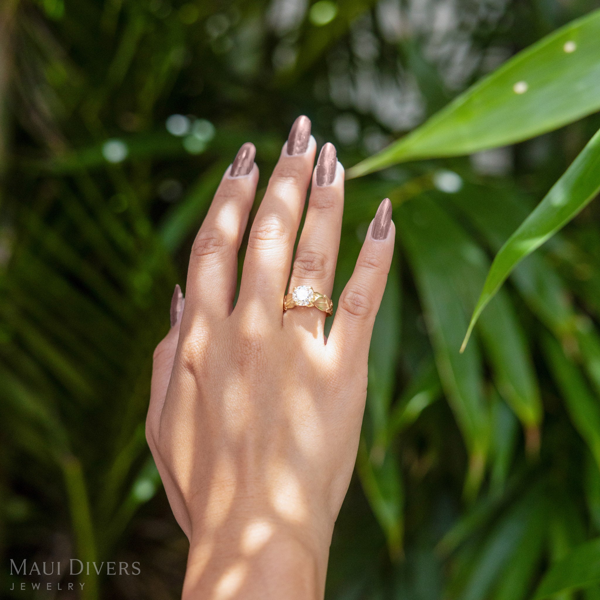 Hand wearing the Maile 2 carat Lab Grown Diamond Engagement Ring in 14k Yellow Gold with a blurry green background