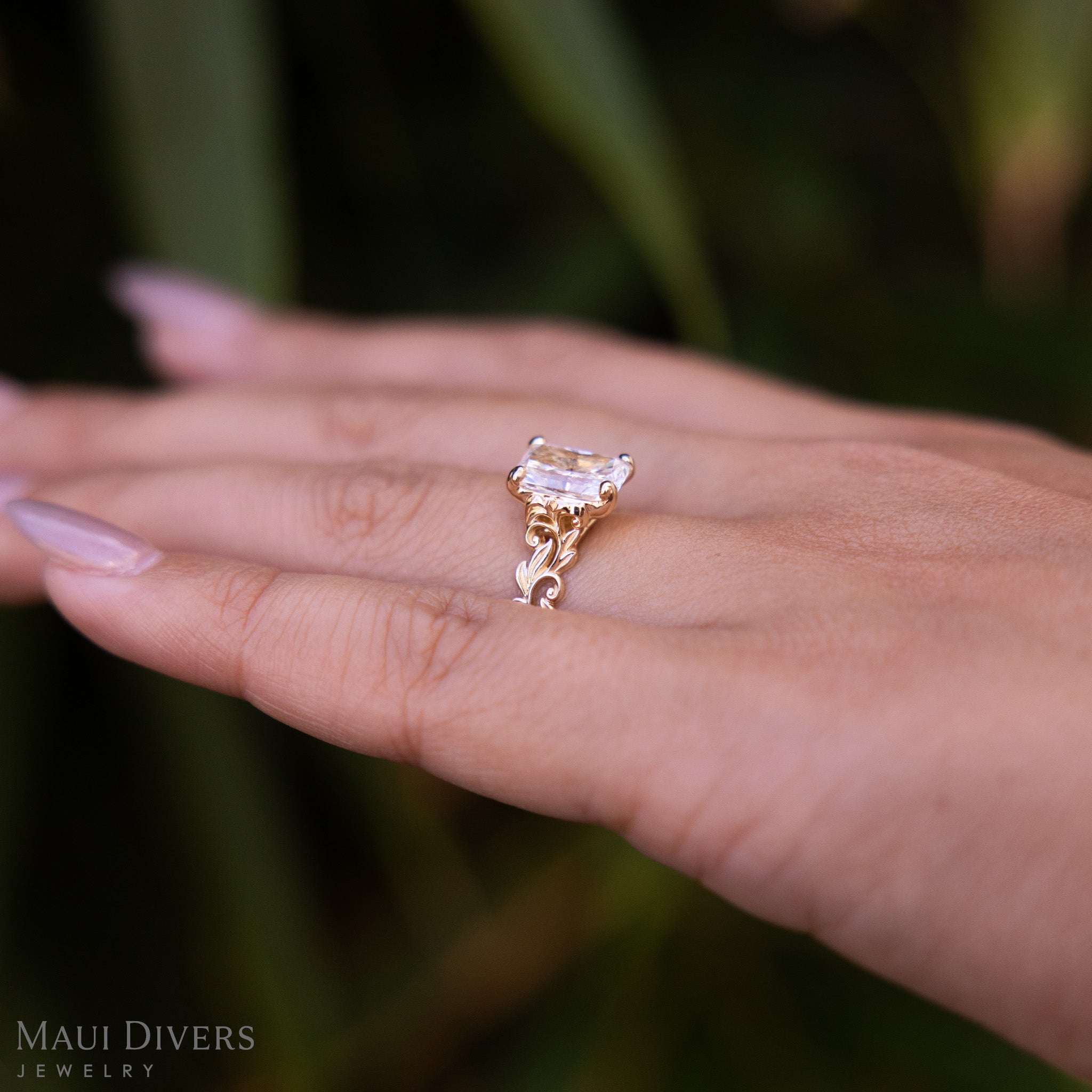 Close-up side view of a Living Heirloom Solitaire Lab Grown Diamond Engagement Ring in 14k yellow gold worn on the ring finger of a hand, against a blurred leafy green background
