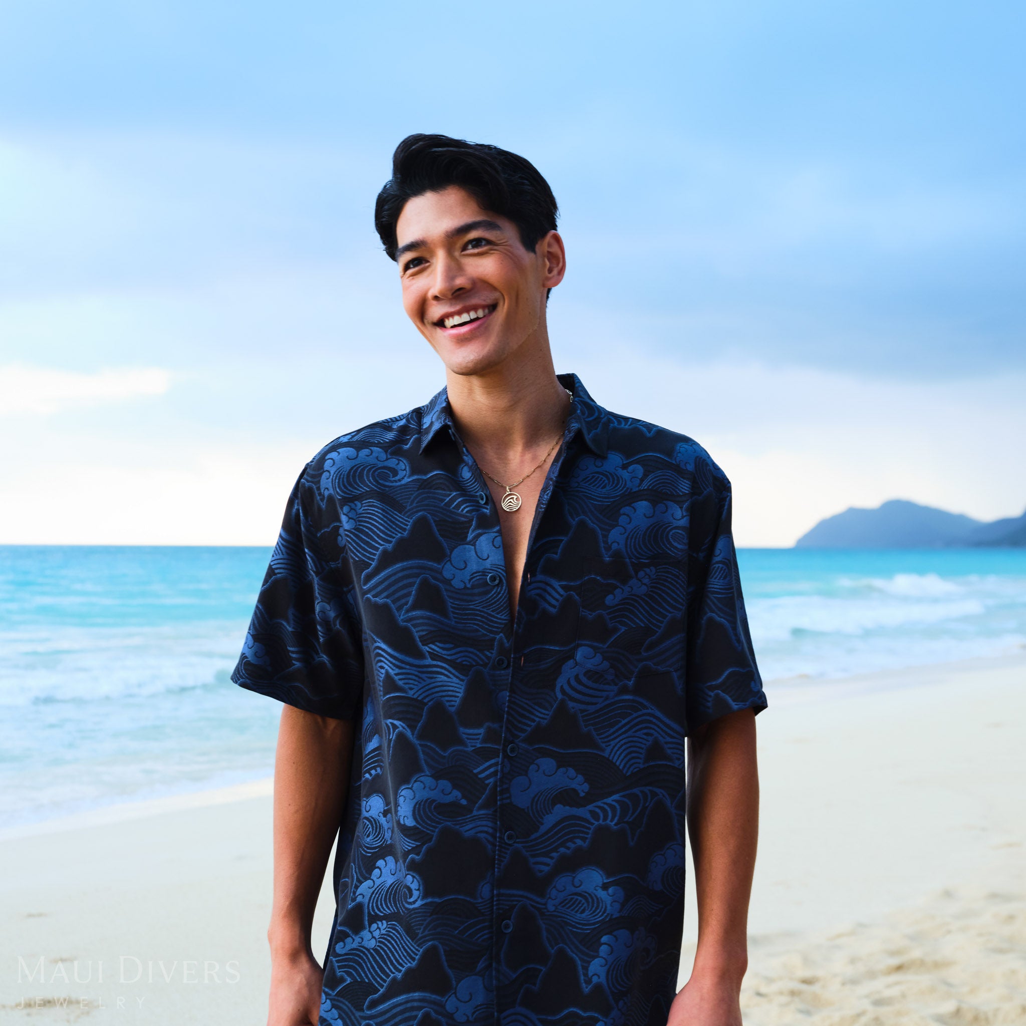 Man wearing a wave pendant on a paperclip chain and a blue patterned shirt on a beach with clear blue sky and ocean.