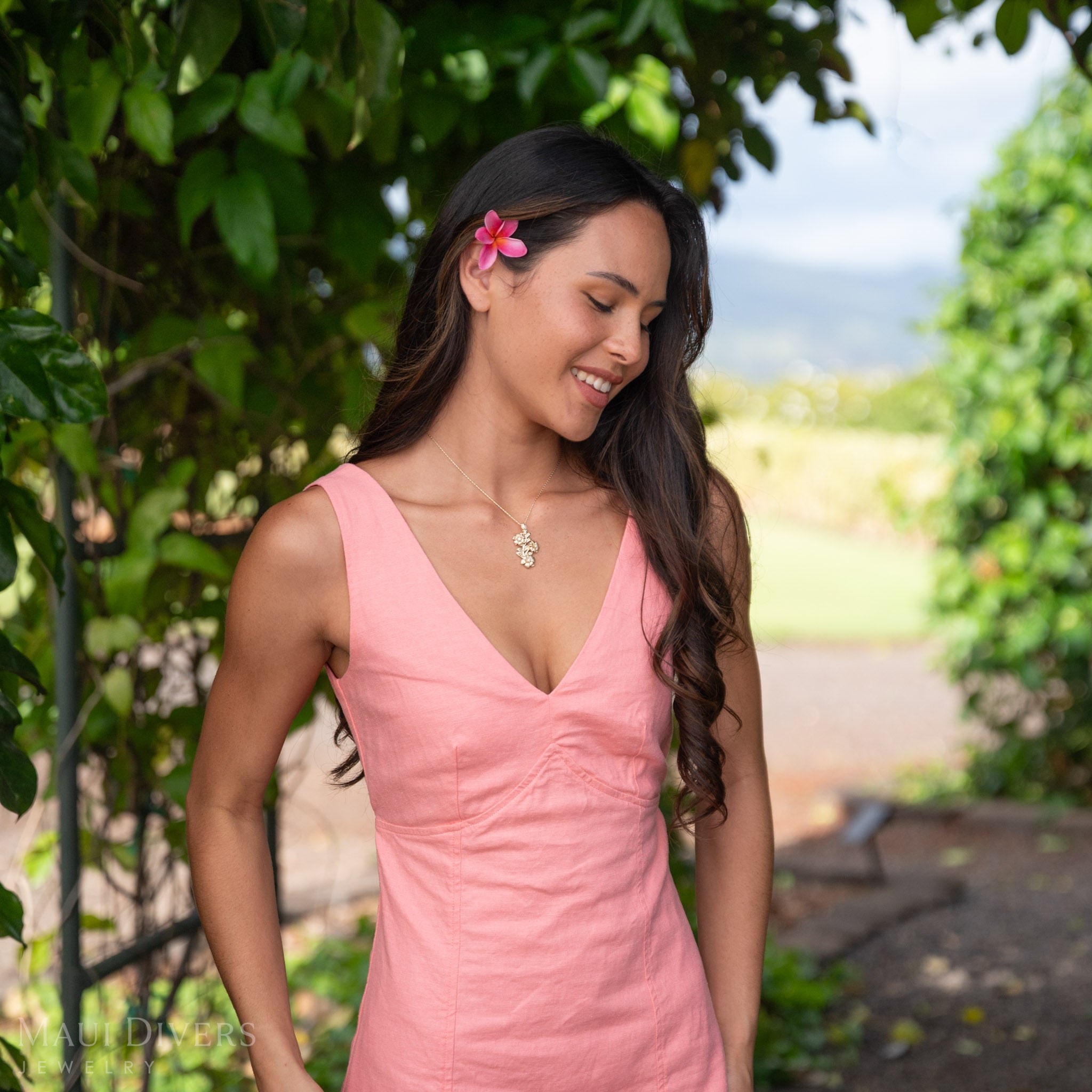 Side-profile of a woman wearing a 14k Gold Hawaiian Gardenia pendant accented with Natural Diamonds with a pink flower in her hair against a green leaf background. 