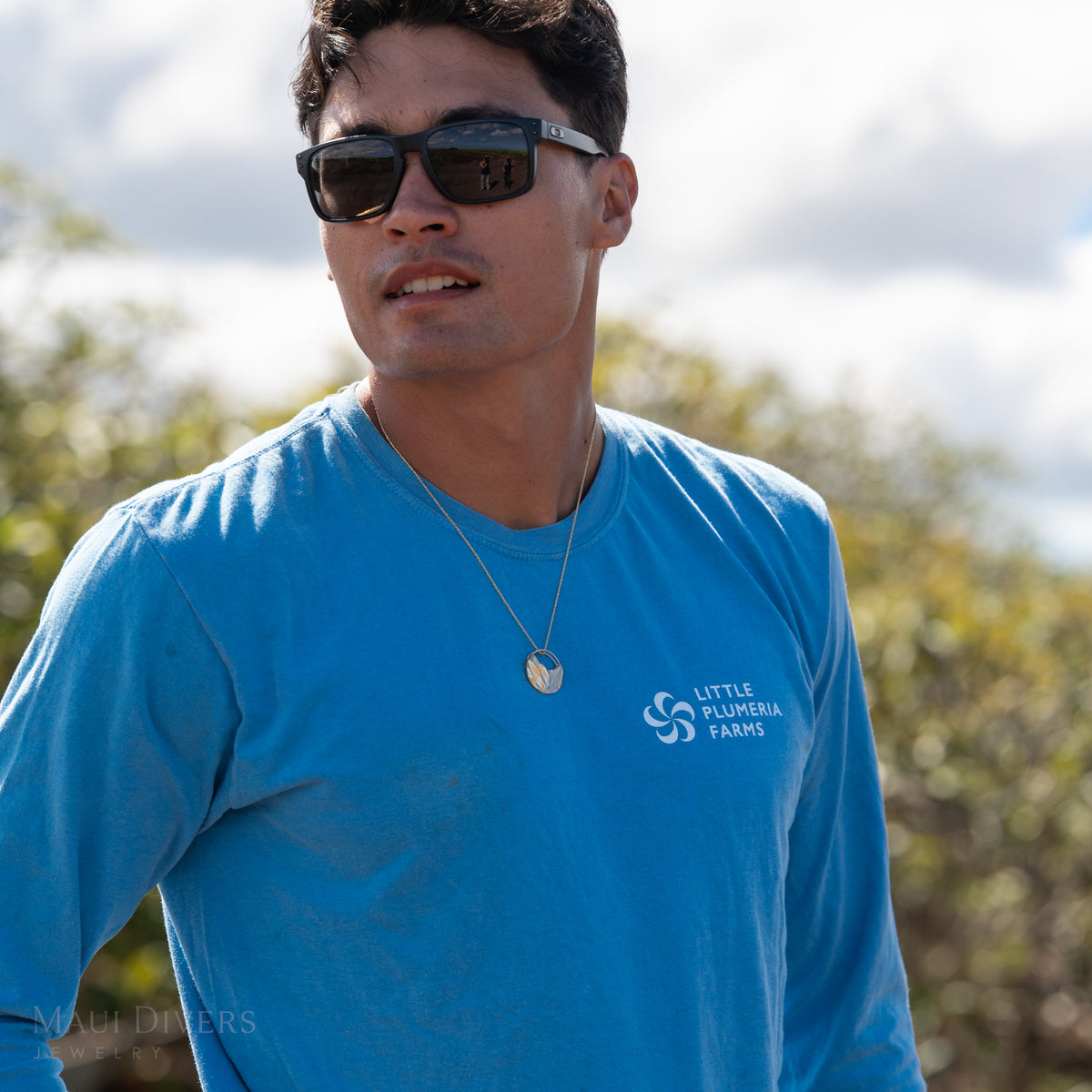 Dane Little in a blue shirt wearing a round pendant with the 14k gold koʻolau mountain range with diamonds, against a blurry cloudy sky and plumeria trees