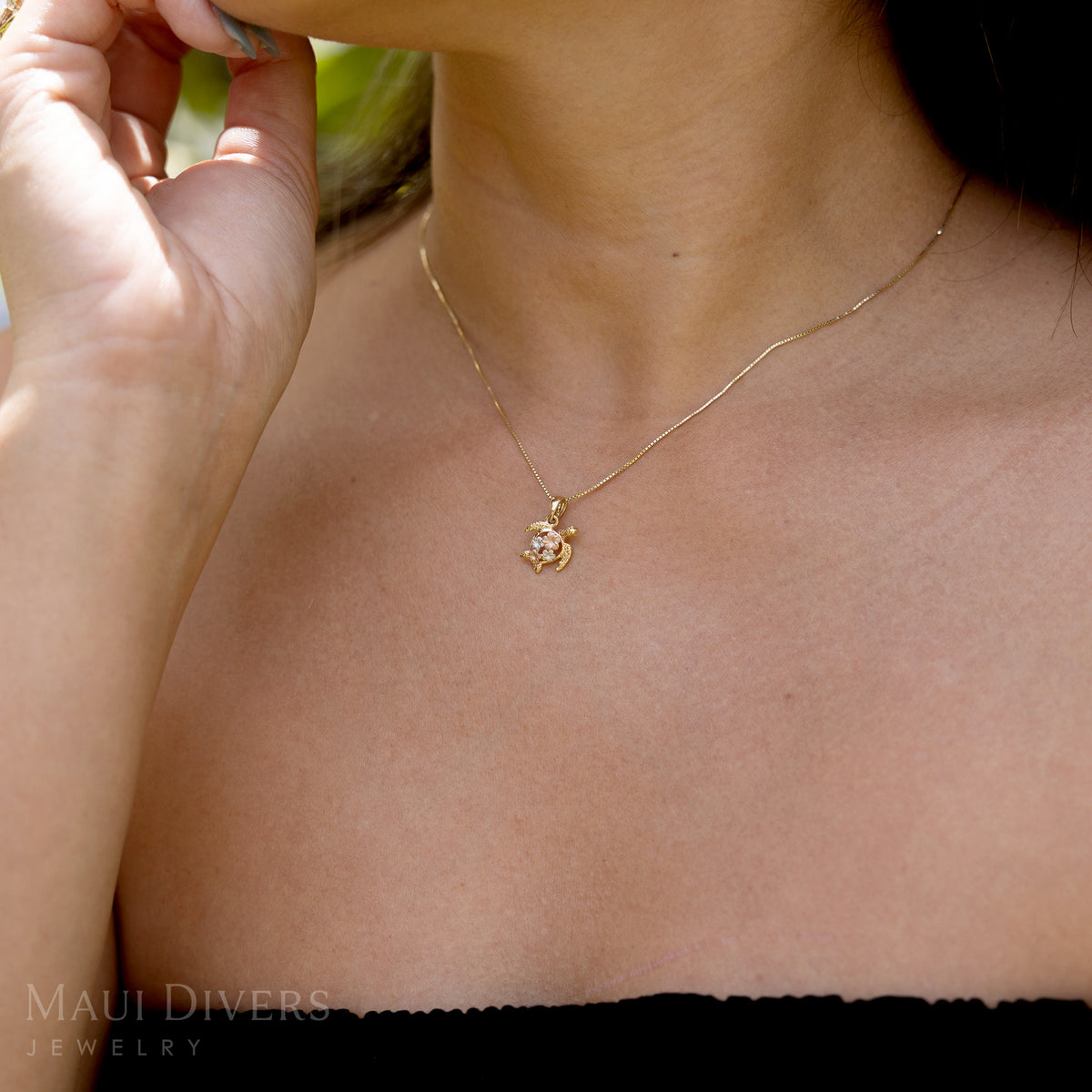 A 14k yellow gold Hawaiian green sea turtle pendant with two rose and two white gold plumerias on its back on a box chain worn around a woman's neck, against a blurred green background