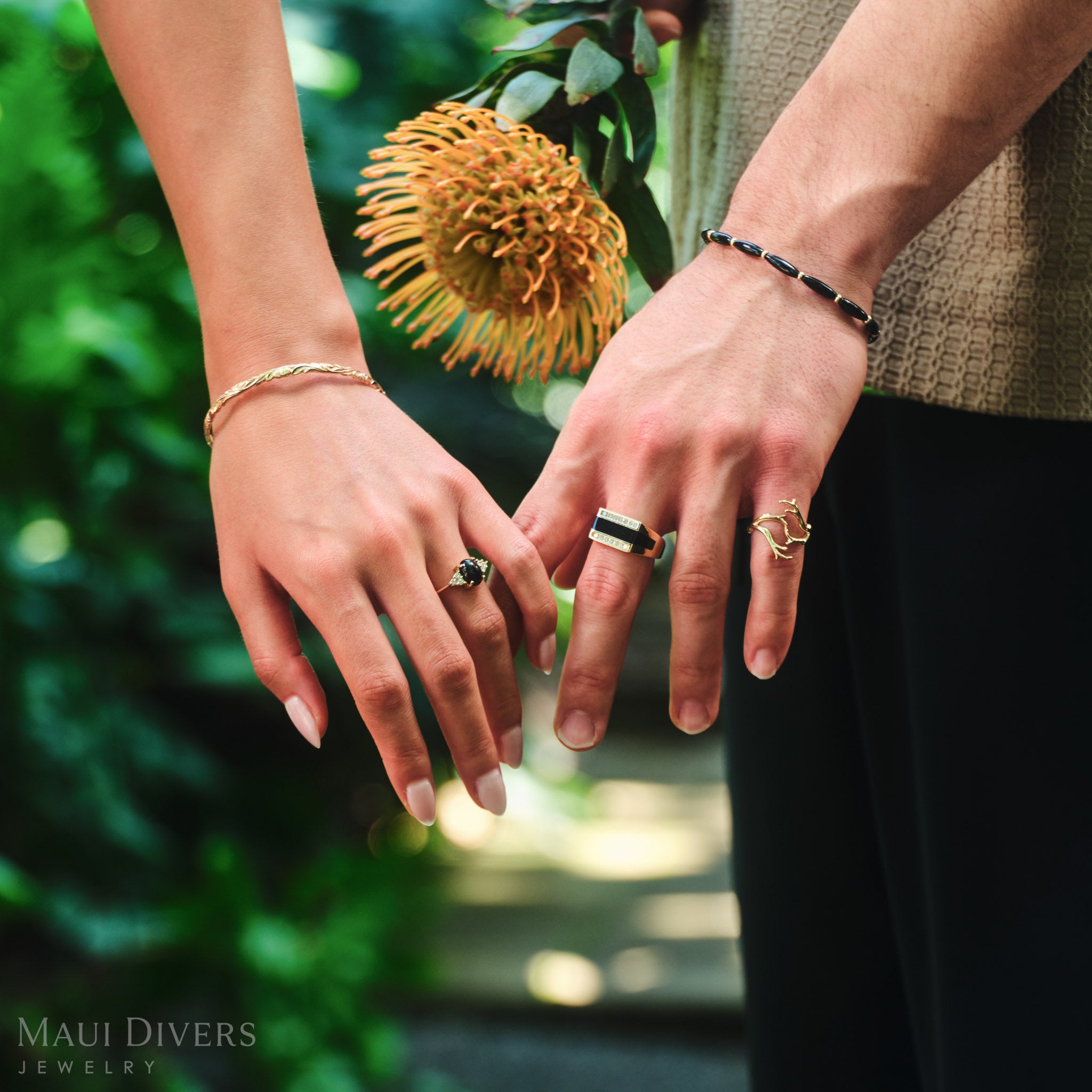 Close-up of a couple's hands, both wearing black coral rings in 14k yellow gold with diamonds, against a blurred forest background