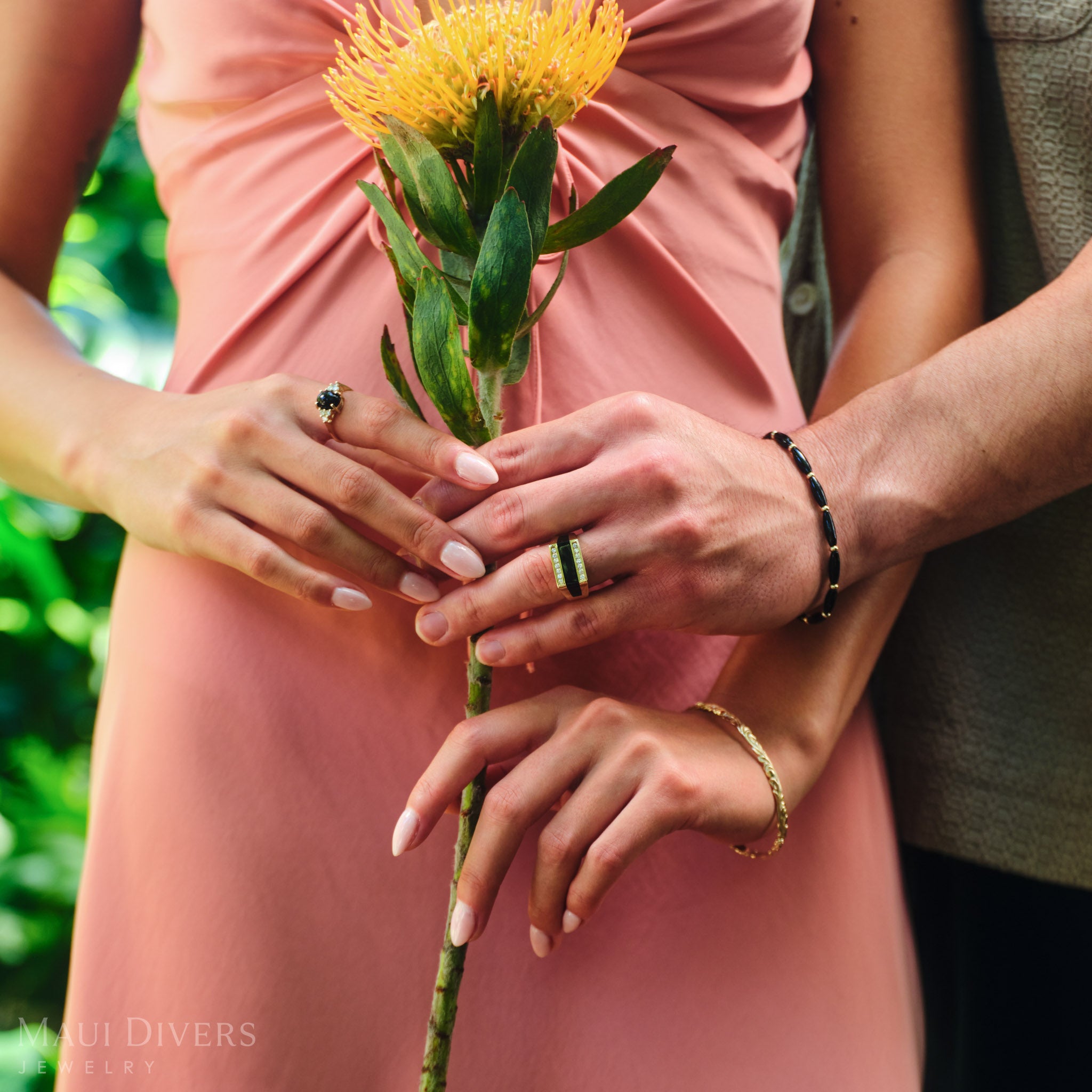 Close-up of a couple's hands holding a flower, both wearing black coral rings in 14k yellow gold with diamonds