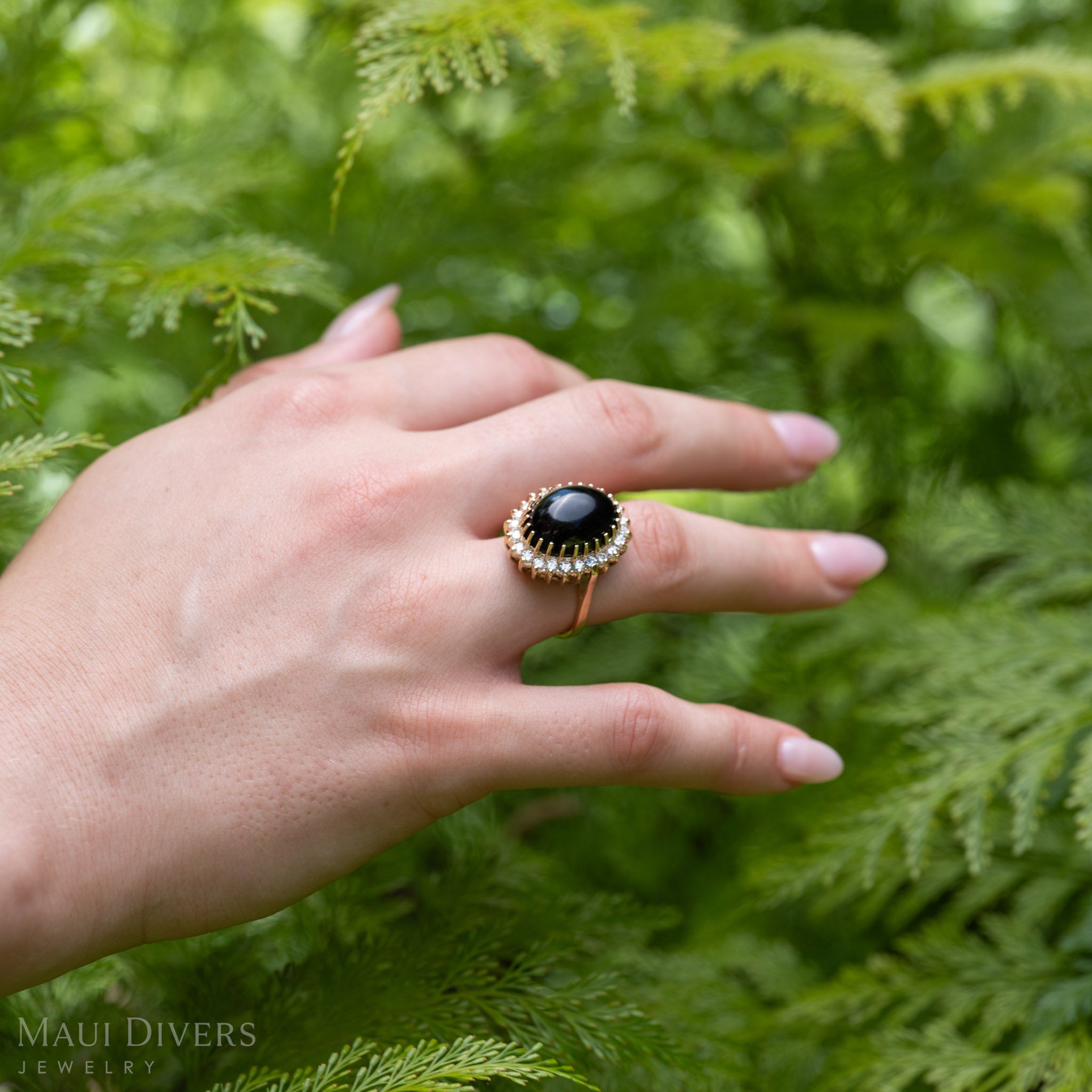 A hand wearing a yellow gold ring with a large oval Hawaiian black coral cabochon and a halo of diamonds, set against a background of green fern foliage. 