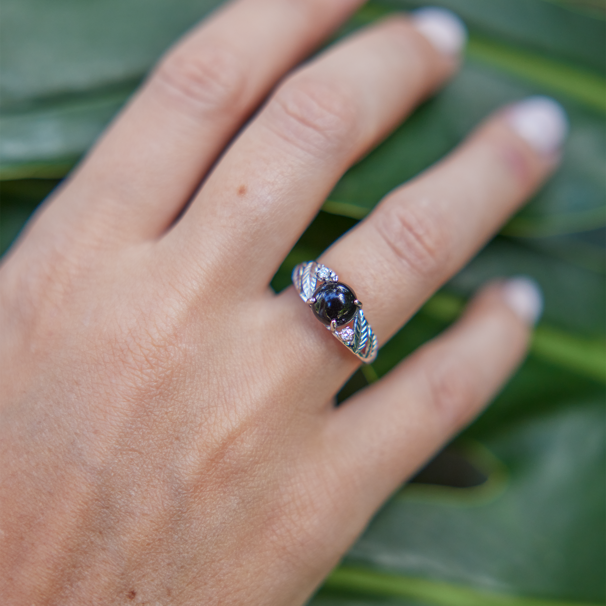 Womanʻs hand wearing Maile Black Coral Ring in White Gold with Diamonds with monstera in the background