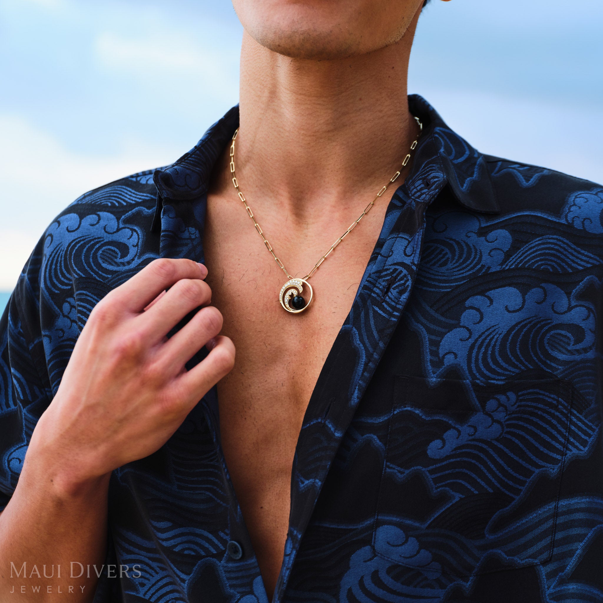 Man wearing a gold necklace with a diamond wave black coral pendant, against a blurred natural background