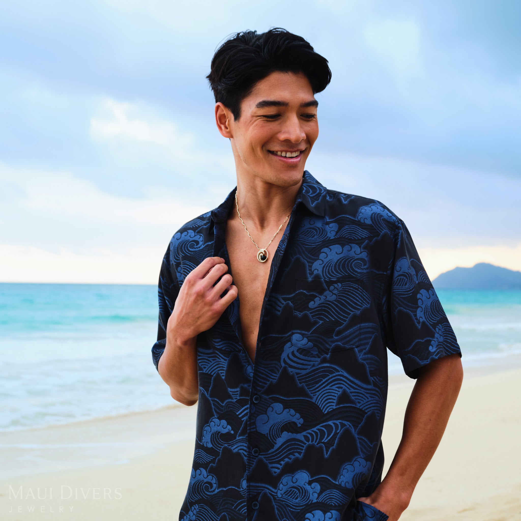 Man wearing a gold necklace with a diamond wave black coral pendant on a beach