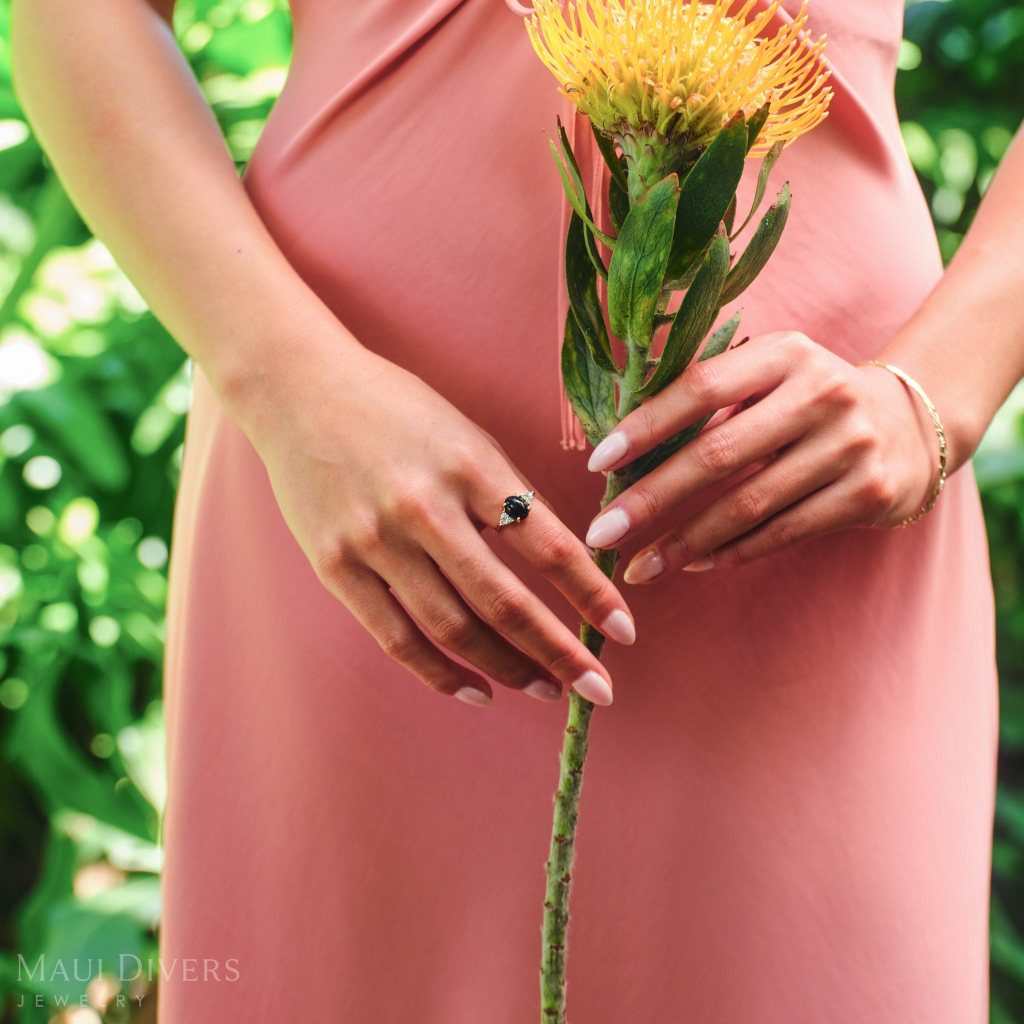 Woman in a pink dress holding a flower wearing a Black Coral Ring in 14k yellow gold with diamonds, against a blurred forest background