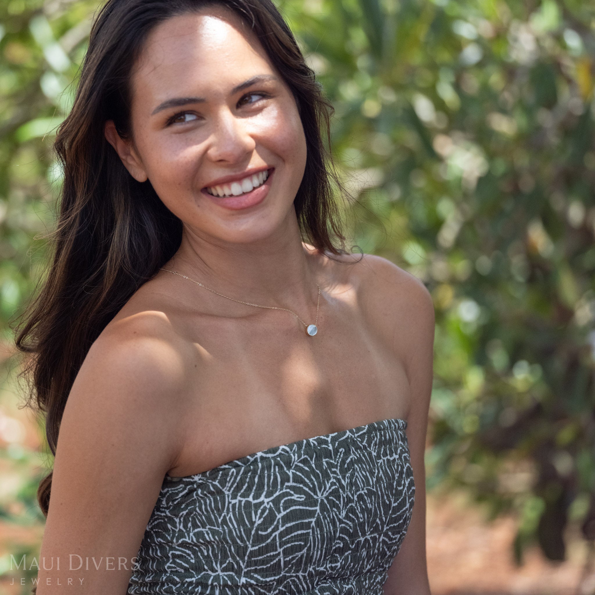 Smiling woman wearing Eclipse Flipside Necklace in 14k Gold showing the Mother of Pearl side against a background of greenery. 