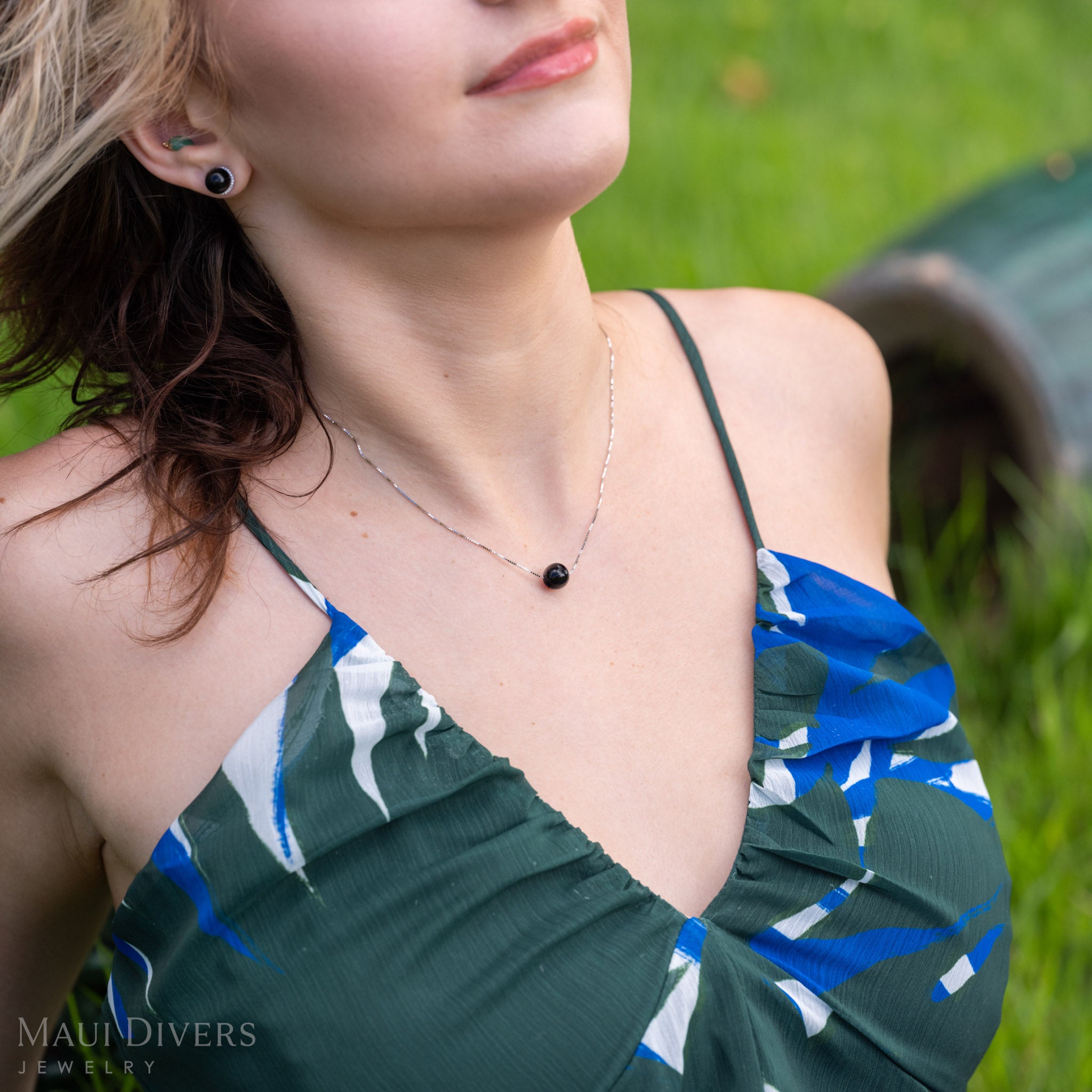 A close-up lifestyle shot of a woman wearing a Hawaiian black coral bead box chain necklace in 14k white gold, paired with a green, blue, and white patterned halter top. She is outdoors on a grassy background.