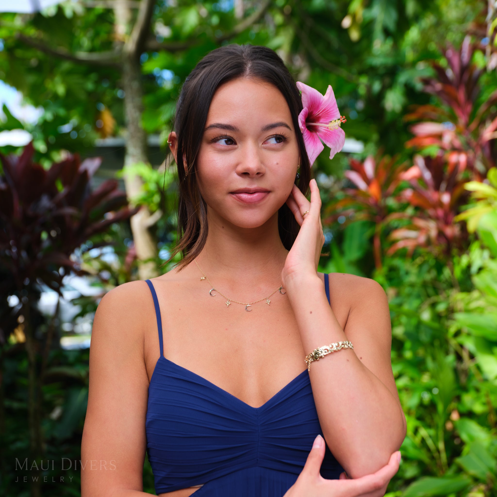 Woman in a blue dress and hibiscus flower in her ear, wearing moon and star necklace in 14k white and yellow gold