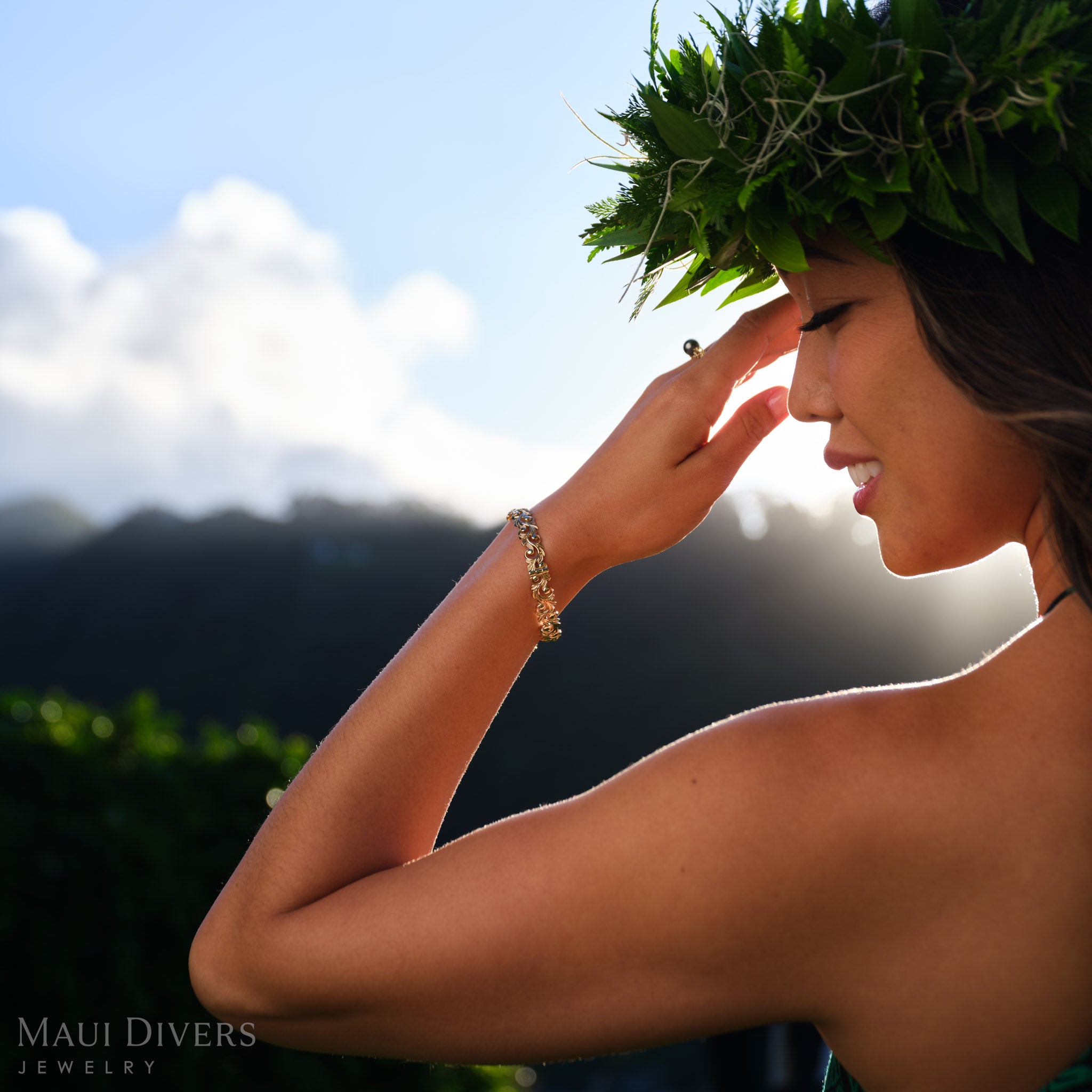 Smiling woman wearing a green leafy headpiece and a Living Heirloom Hinge Bracelet in 14k yellow gold on her left wrist, against a blurred mountain and sky background