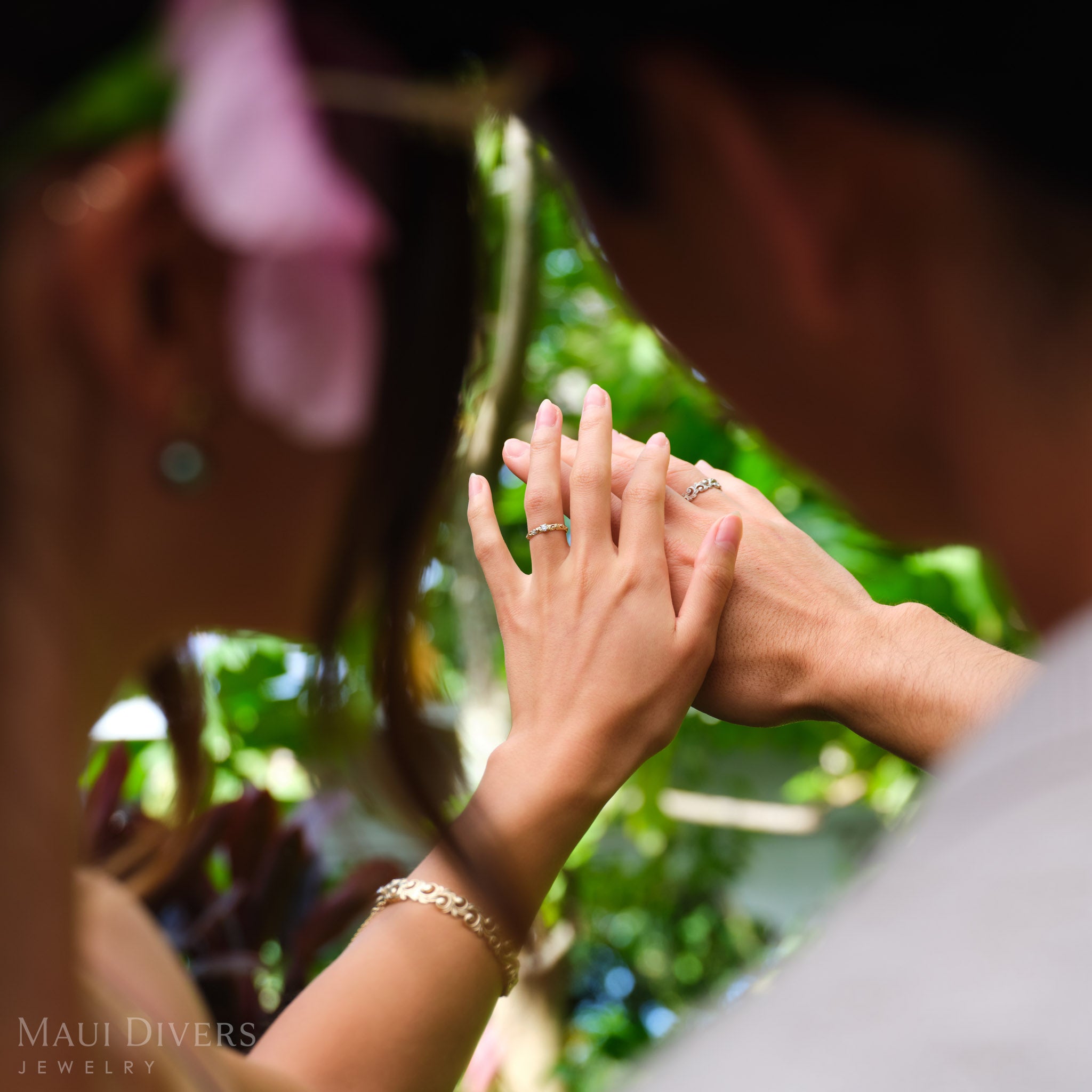 Two people's heads side by side, looking down at their joined hands, with a woman wearing a Living Heirloom Ring in 14k yellow gold with a diamond on her ring finger, against a blurred tropical background