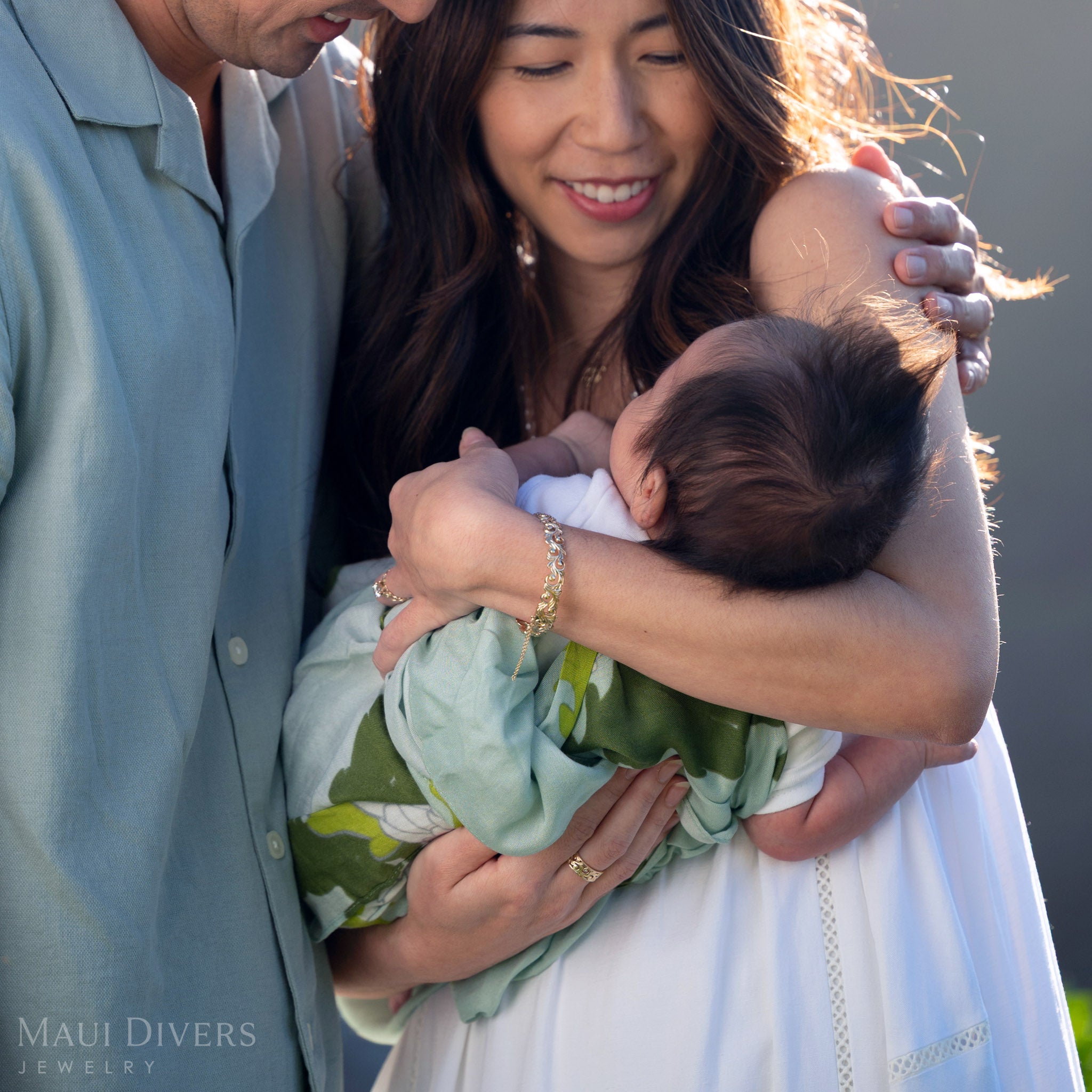 Smiling woman wearing a Living Heirloom Hinge Bracelet in 14k yellow gold on her left wrist, with her arm wrapped around a baby and another person's arms wrapped around her