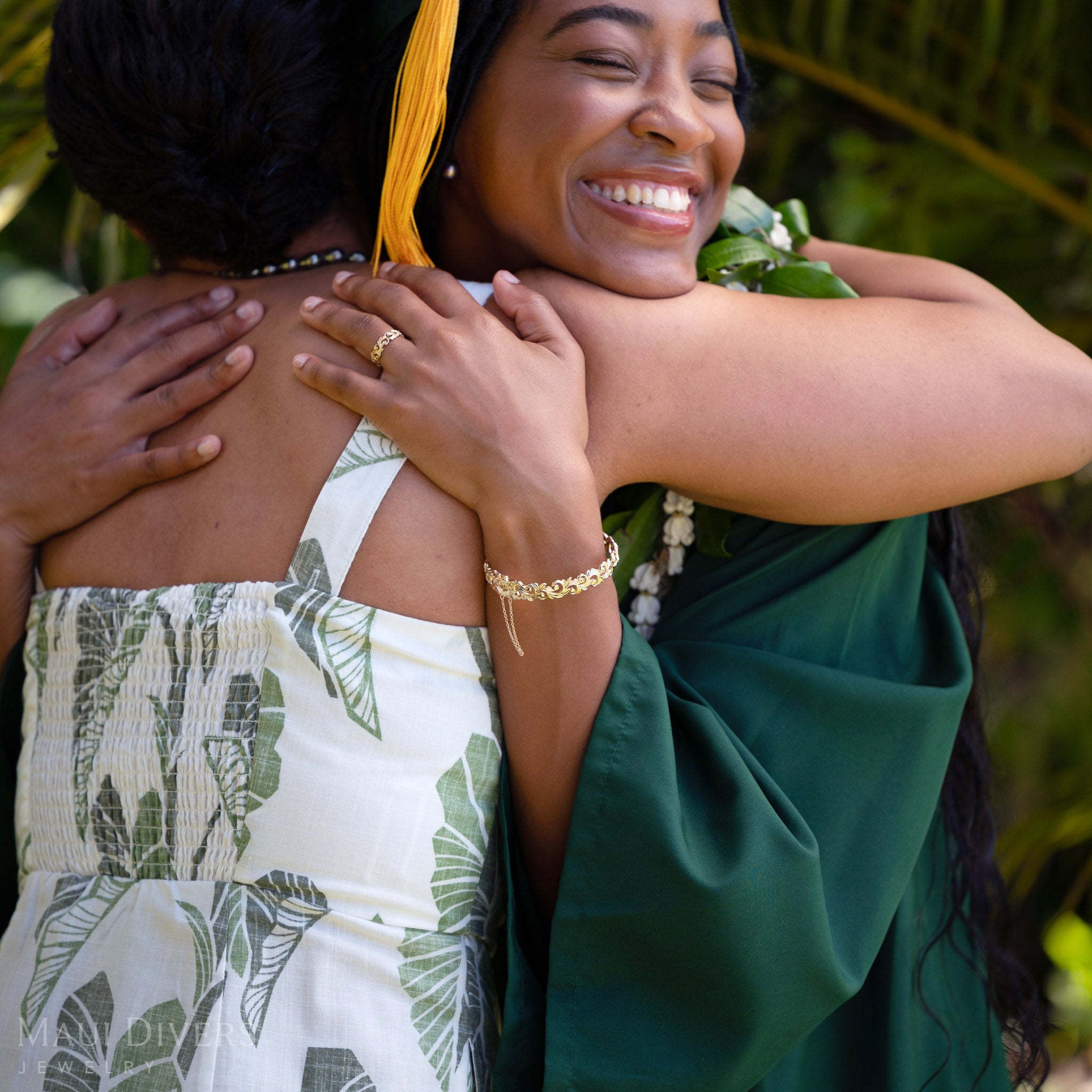 Smiling woman wearing a Living Heirloom Hinge Bracelet in 14k yellow gold on her left wrist, embracing another woman against a blurred green background