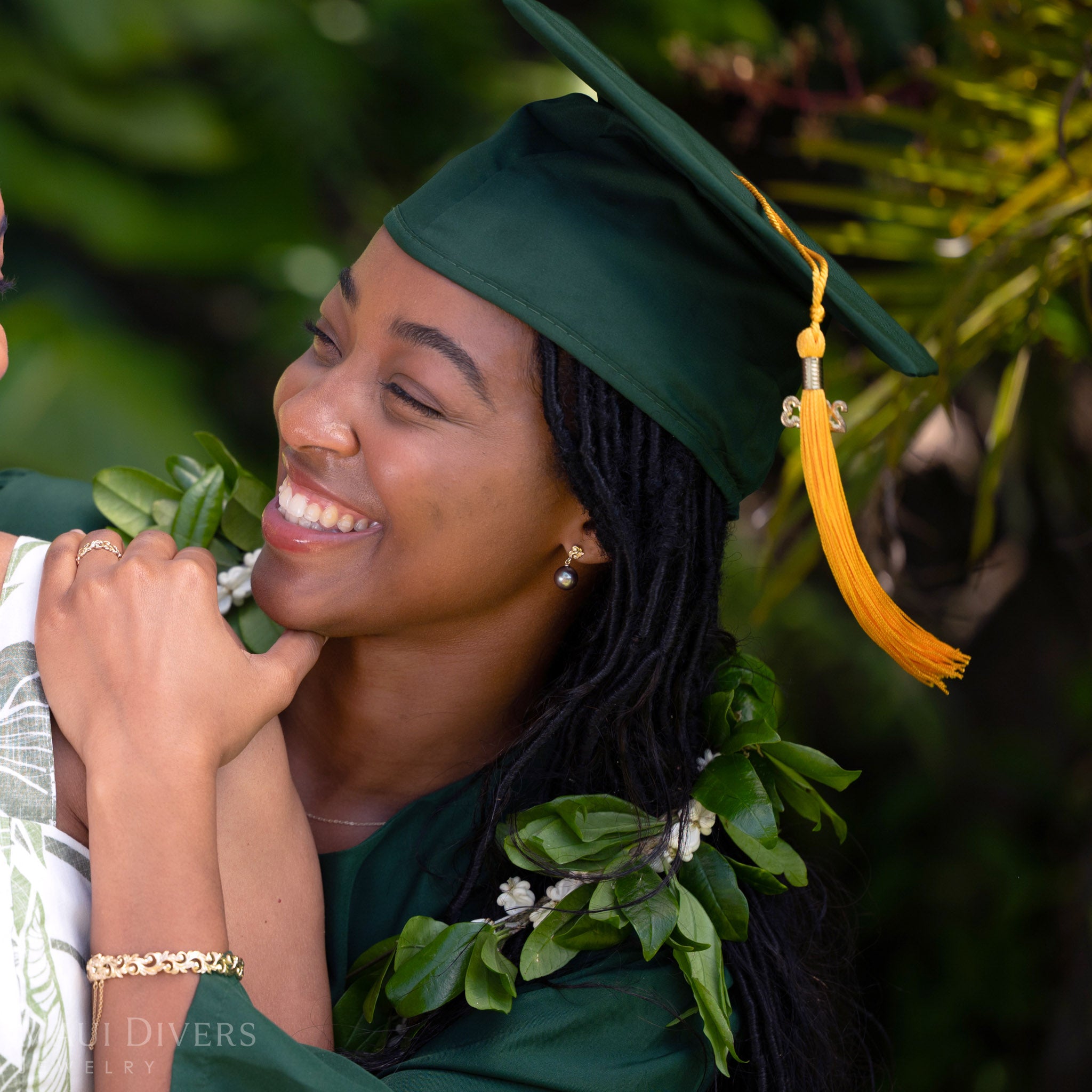 Smiling woman in a green graduation cap and gown wearing a Living Heirloom Freshwater Pearl Earring in 14k yellow gold with diamond accents on her left ear, looking at another person against a blurred tropical background